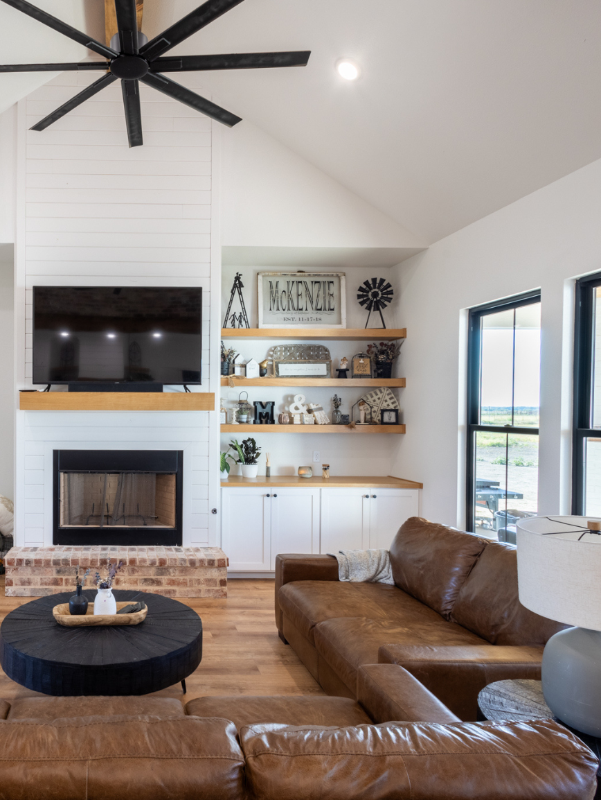 Living room with a brown leather sofa, a round black coffee table with decorative items, a white fireplace with a mounted TV above, wooden shelves with decor, a window showing an outdoor view, and a ceiling fan.