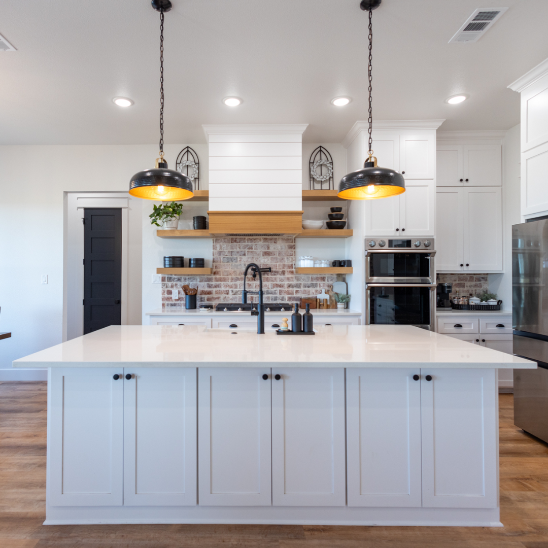 Modern kitchen with white cabinets, a large white island, black pendant lights, an exposed brick backsplash, and stainless steel appliances.