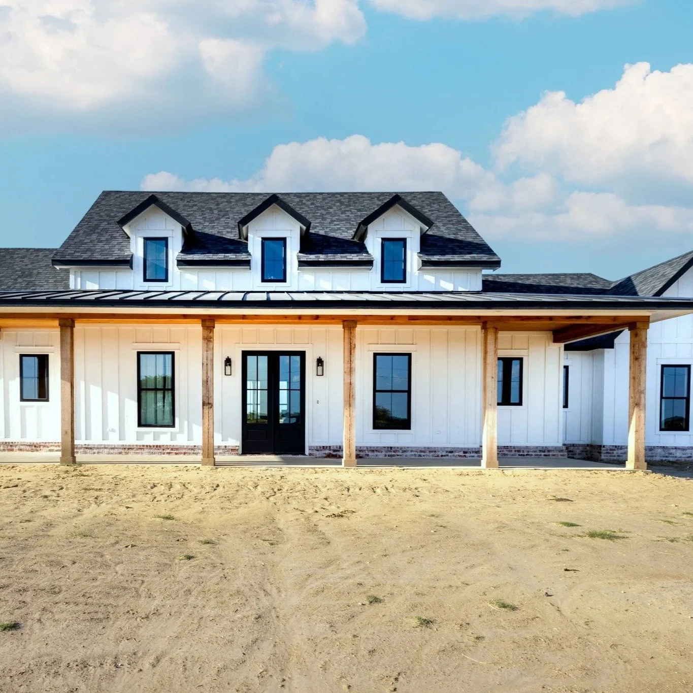 Front view of a modern house with white siding, black framed windows and a black front door under a covered porch supported by wooden pillars, with a partly cloudy sky in the background and sandy ground in the foreground.