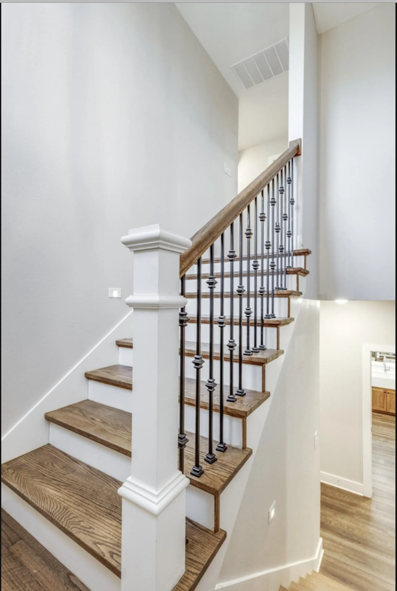 Interior view of a wooden staircase with black metal balusters and white newel posts, leading upstairs in a home with white walls and hardwood flooring.