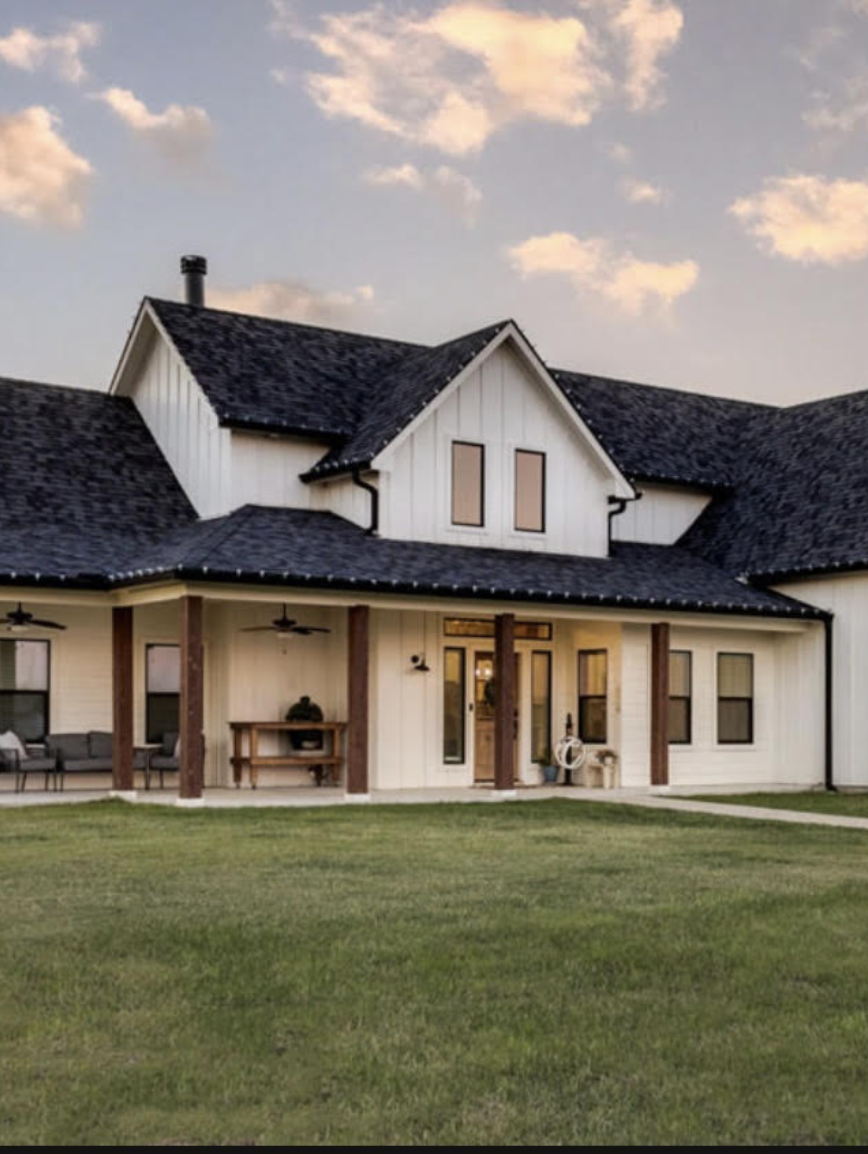 A two-story house with a dark shingled roof, white siding, and a front porch with furniture and decor, set against a partly cloudy sky.