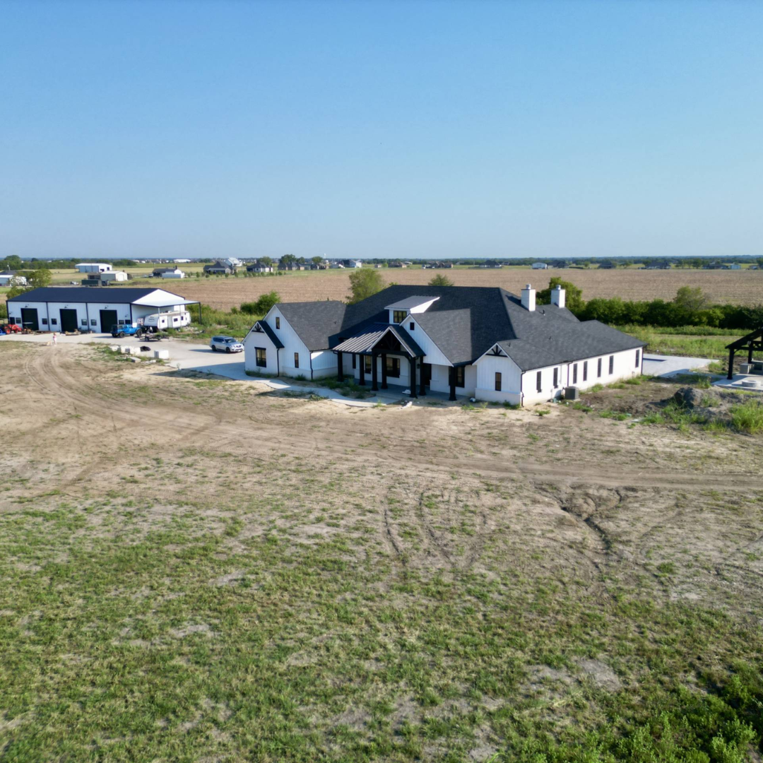 A house under construction in a rural area with a large open field, a clear blue sky, and construction vehicles nearby.