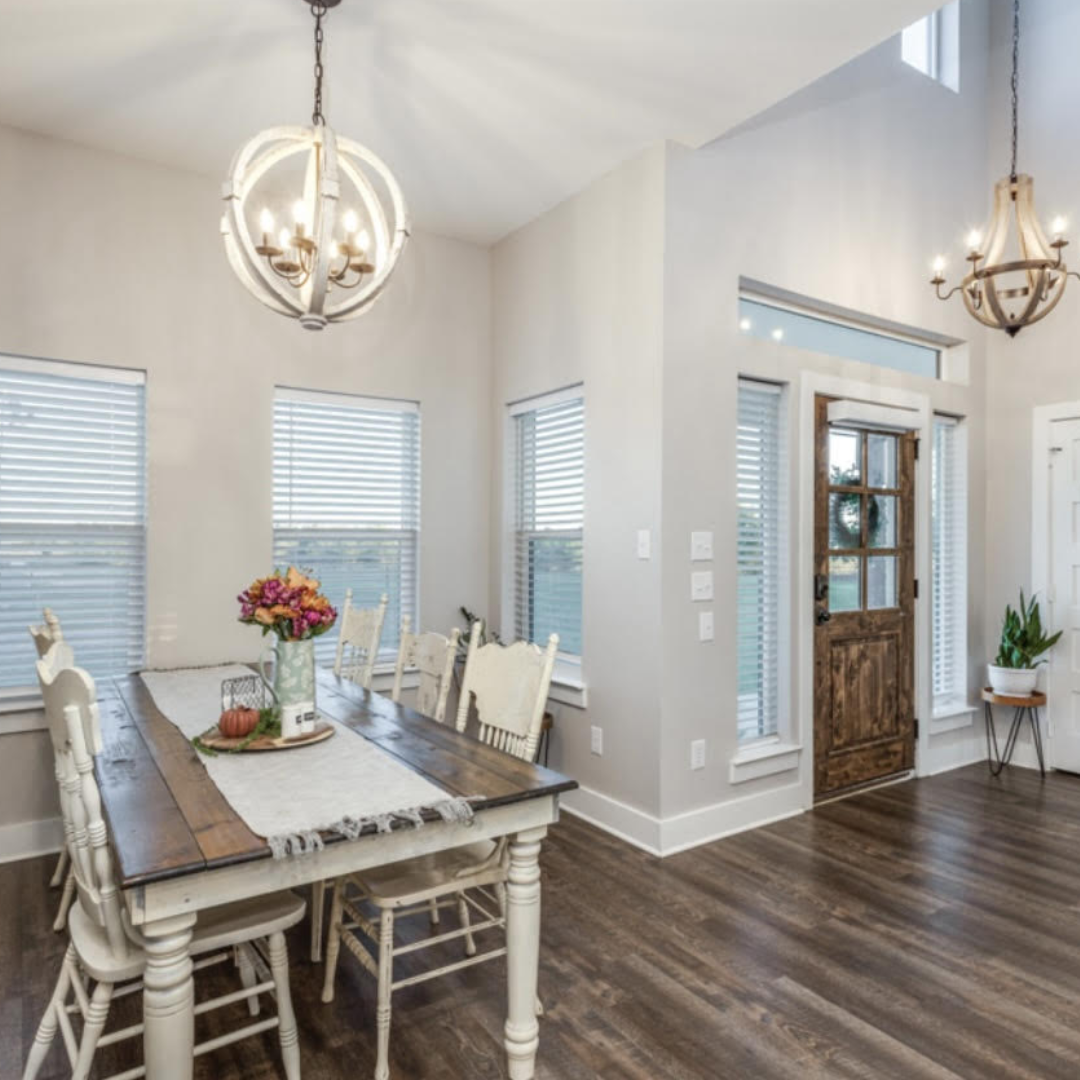 Dining room with a wooden table, white chairs, window blinds, and two hanging chandeliers. Decor includes a vase of flowers and a pumpkin on the table, with hardwood flooring and a wooden front door.