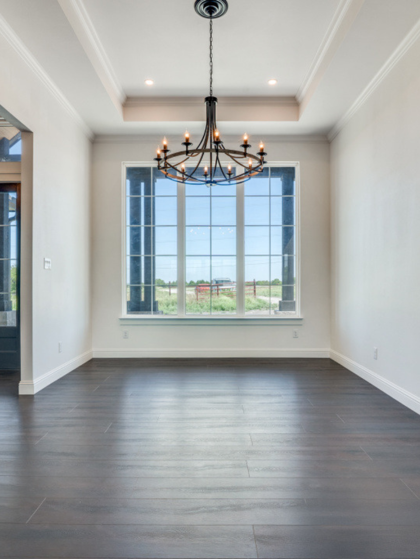 Empty dining room with dark wood flooring, white walls, large window, and black chandelier.
