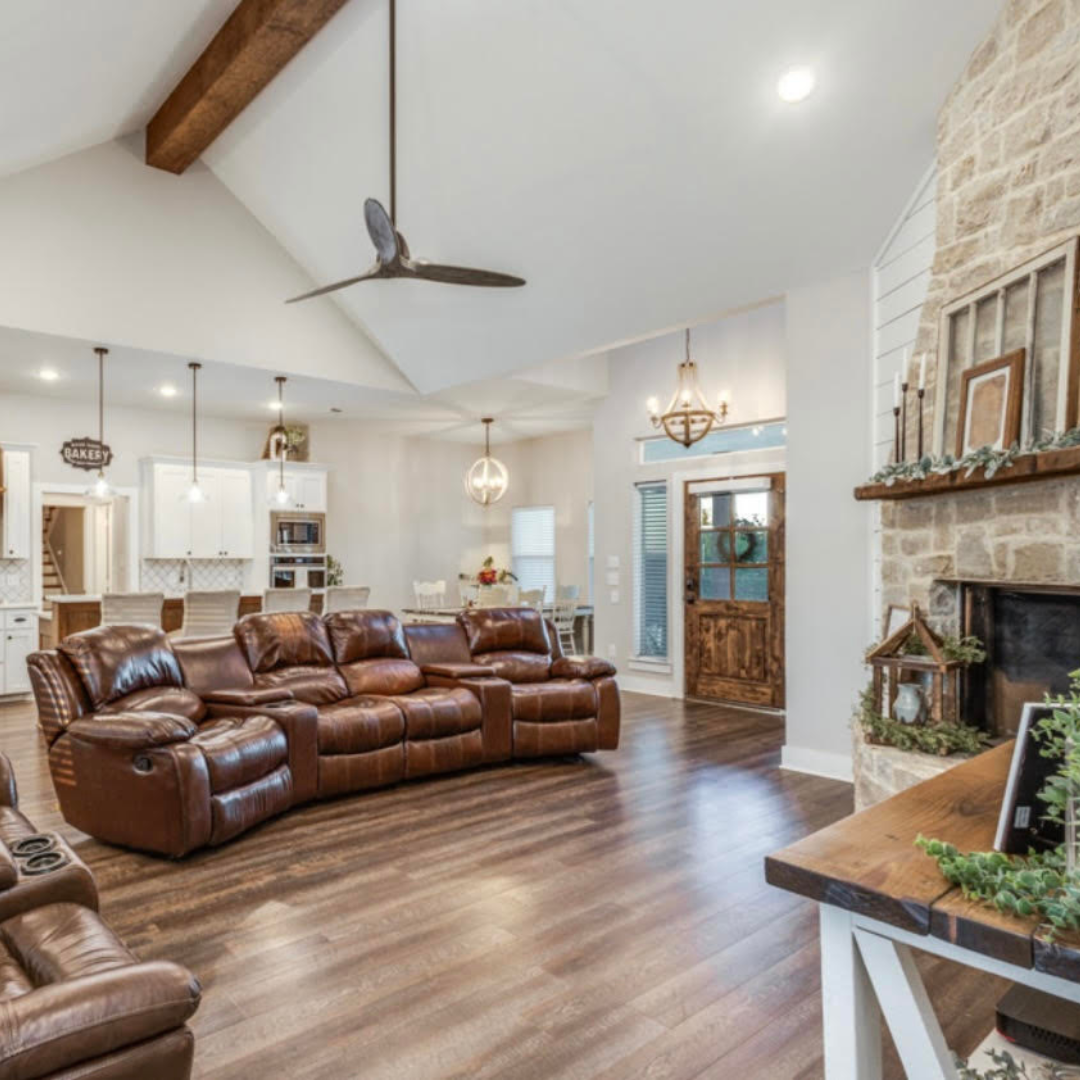 A spacious living room with brown leather sofas, hardwood floors, a stone fireplace decorated with greenery, a wooden front door with a wreath, and bright lighting from ceiling fixtures and windows.