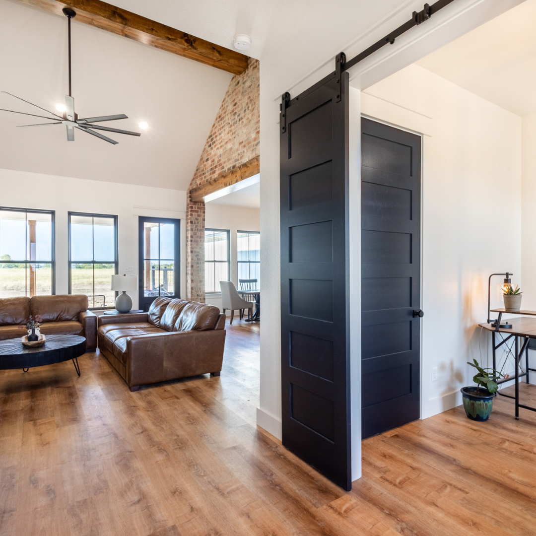 Living room with leather sofa, wooden floor, large windows, brick wall, wooden beams, and a black sliding barn door.