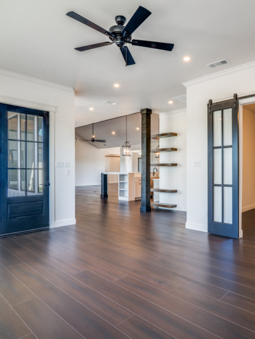 Empty room with dark wood flooring, white walls, black ceiling fans, and open kitchen in background with wooden shelves and black beams.