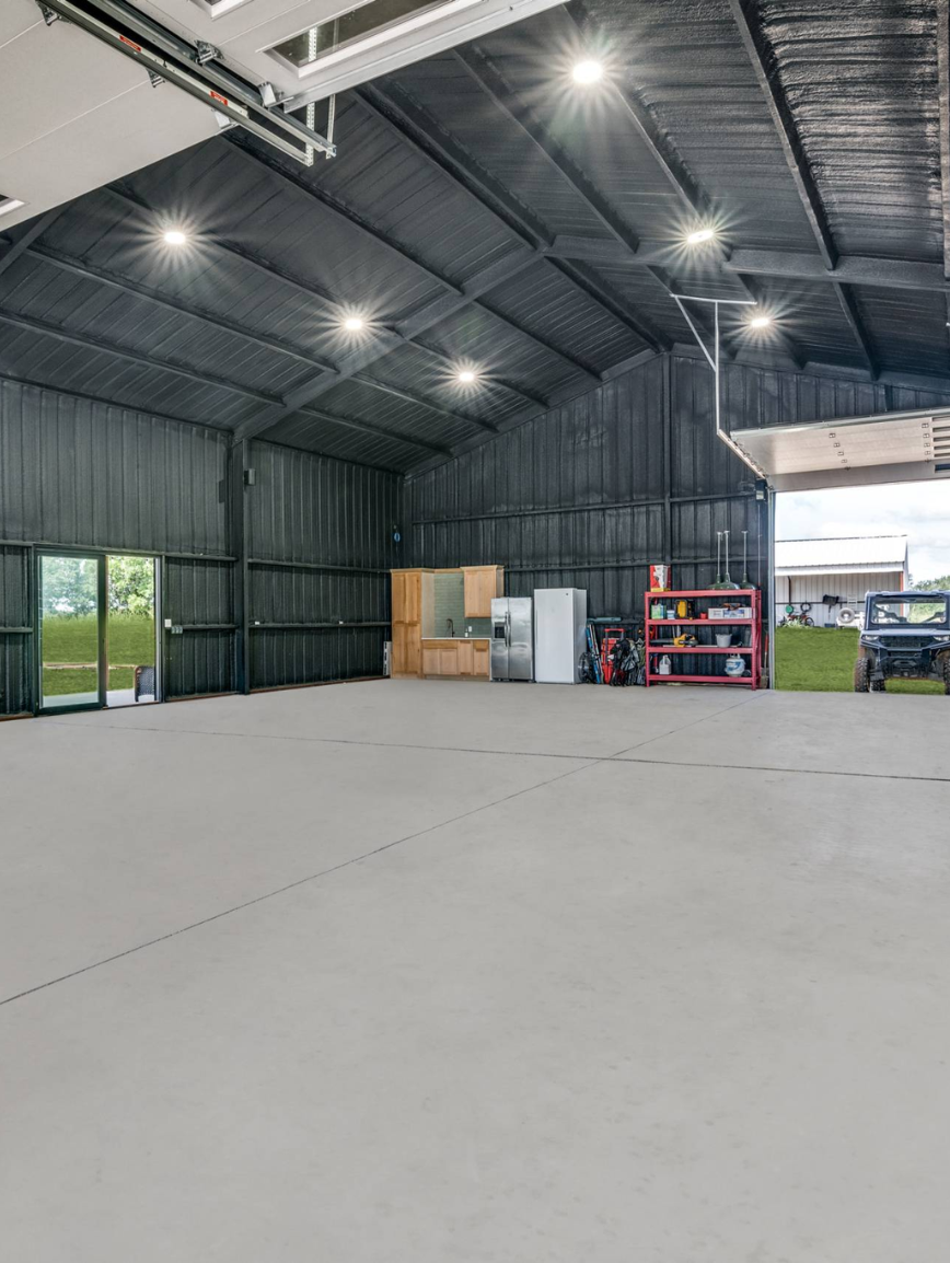 Empty metal storage building with concrete floor, lighting, and shelves with tools and equipment near the back wall, open garage door showing grass and building outside.