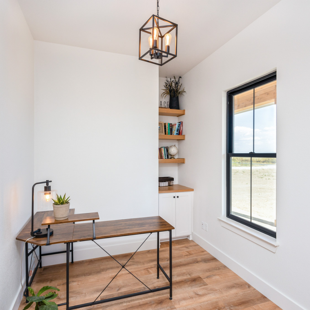 A small workspace with a wooden desk, a potted plant, and a modern black desk lamp in a brightly lit room with white walls, wooden flooring, a window showing an outdoor landscape, and built-in shelves with books and decor.