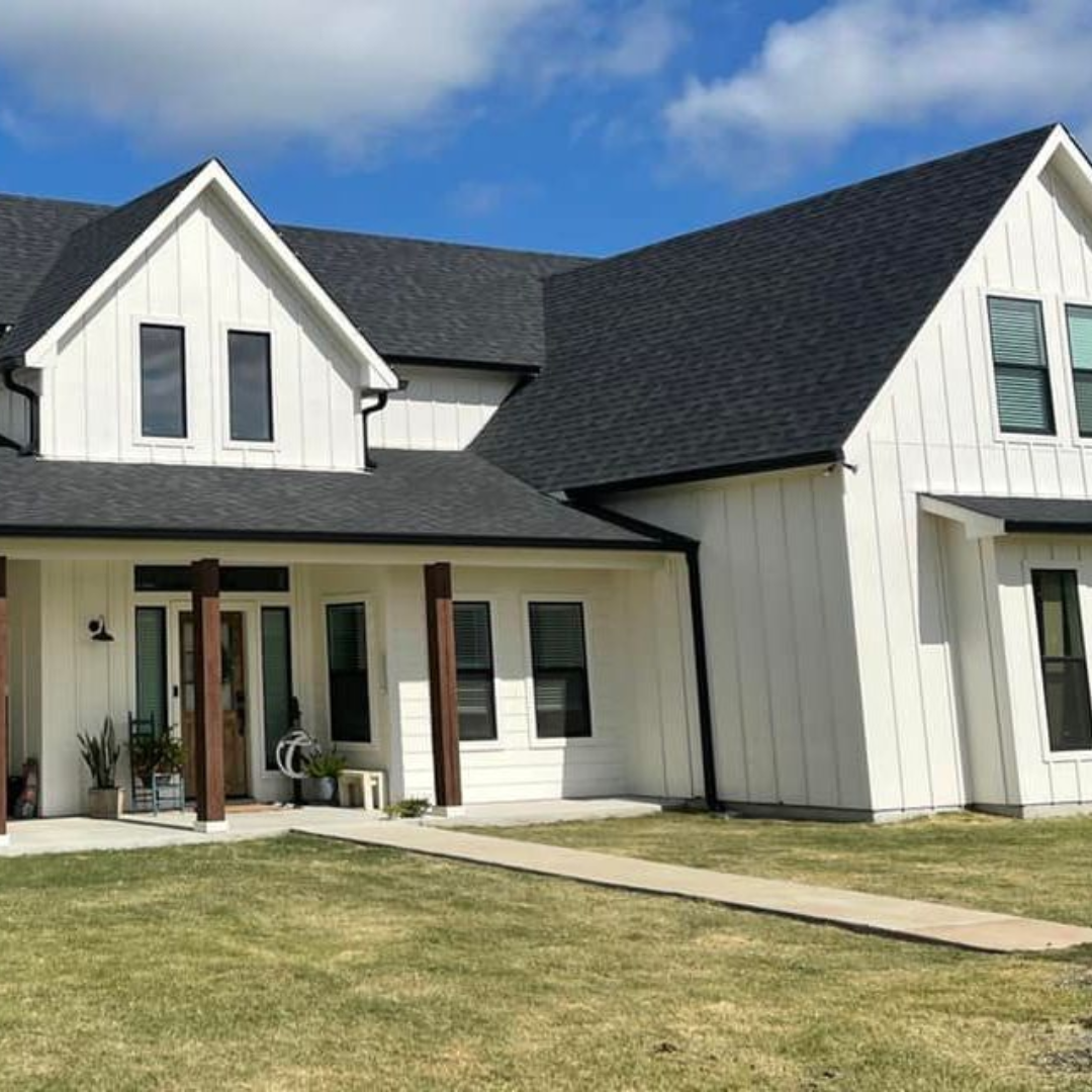 Front view of a modern white house with black roof shingles, multiple gables, and windows, under a blue sky with some clouds.