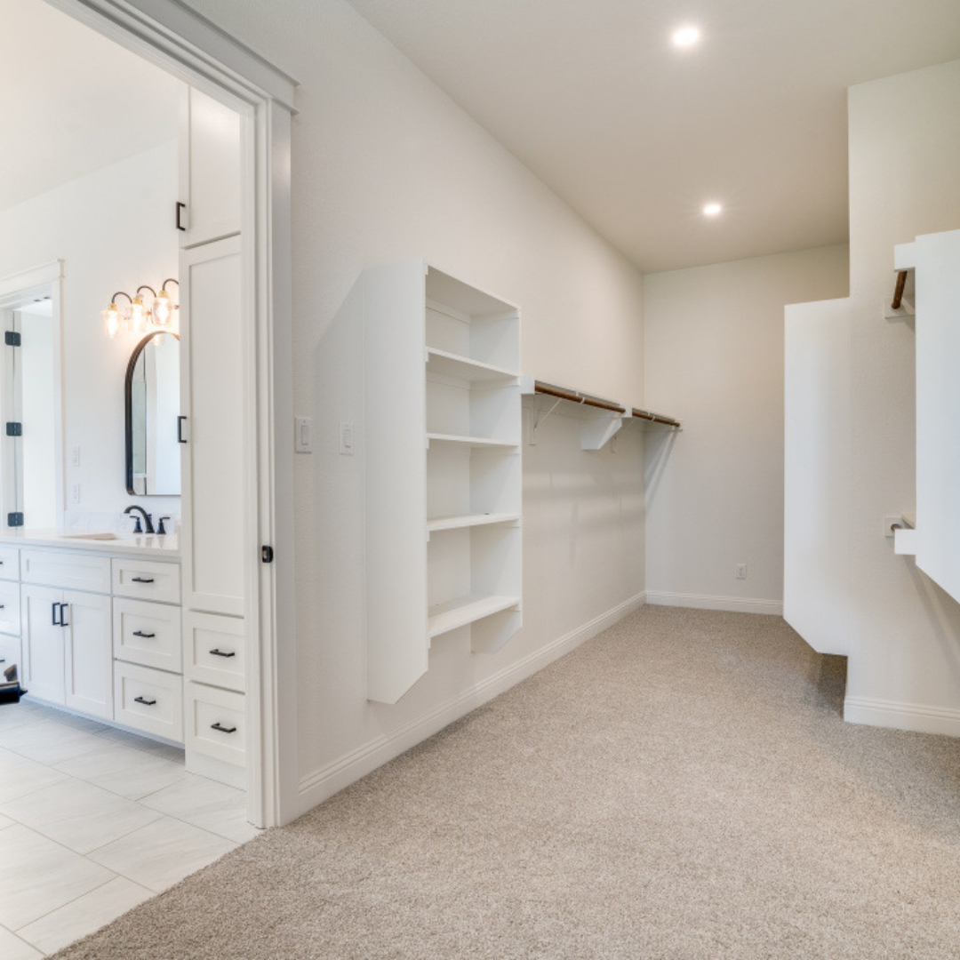Empty walk-in closet with white shelving and beige carpet, adjacent to a bathroom with white cabinetry and black hardware.