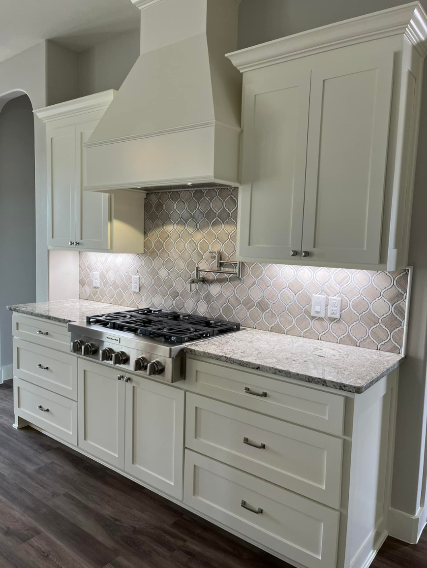 White kitchen cabinets and countertop with a gas stove, backsplash tiles, and a wall-mounted small shelf, with gray walls and dark wood flooring.