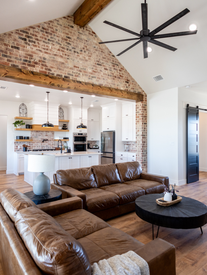 Living room with exposed brick wall, two leather sofas, round black coffee table with decorative items, and a dining area with hanging pendant lights in the background. The ceiling is vaulted with a large black ceiling fan.