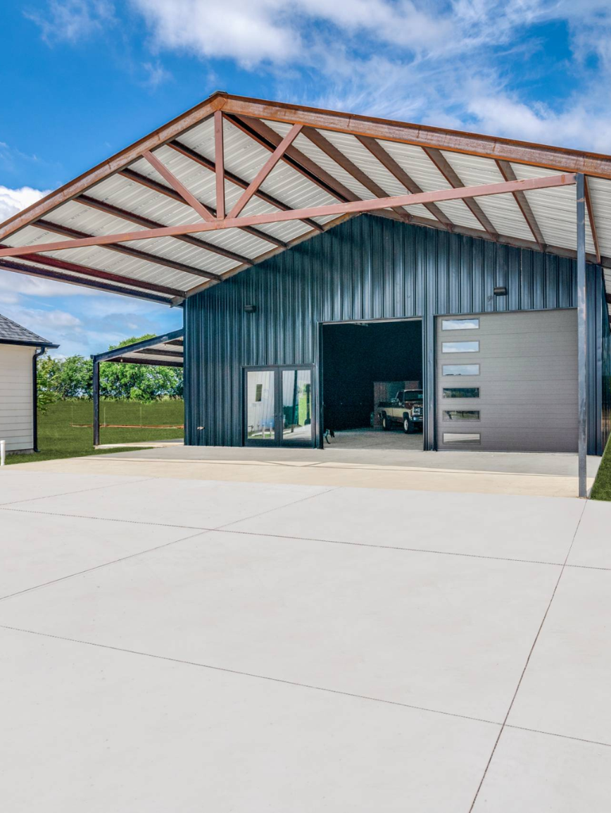 Large modern garage with metal siding, open door revealing a vehicle inside, concrete driveway, metal roof with exposed wooden trusses, and a small side building, under a partly cloudy sky.