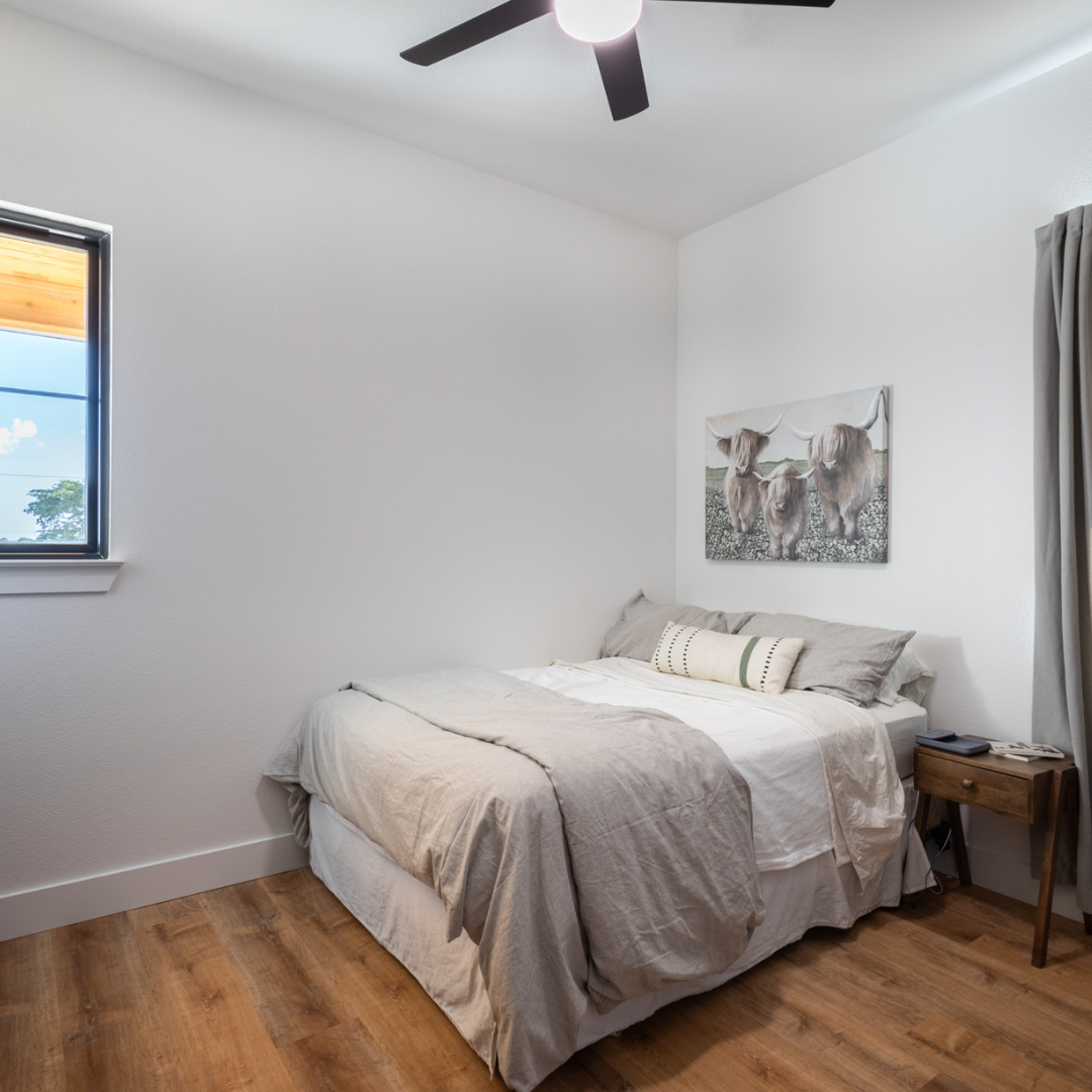 A minimalist bedroom with a bed, painting of two cows, wooden nightstand with phone, gray curtains, and a ceiling fan.