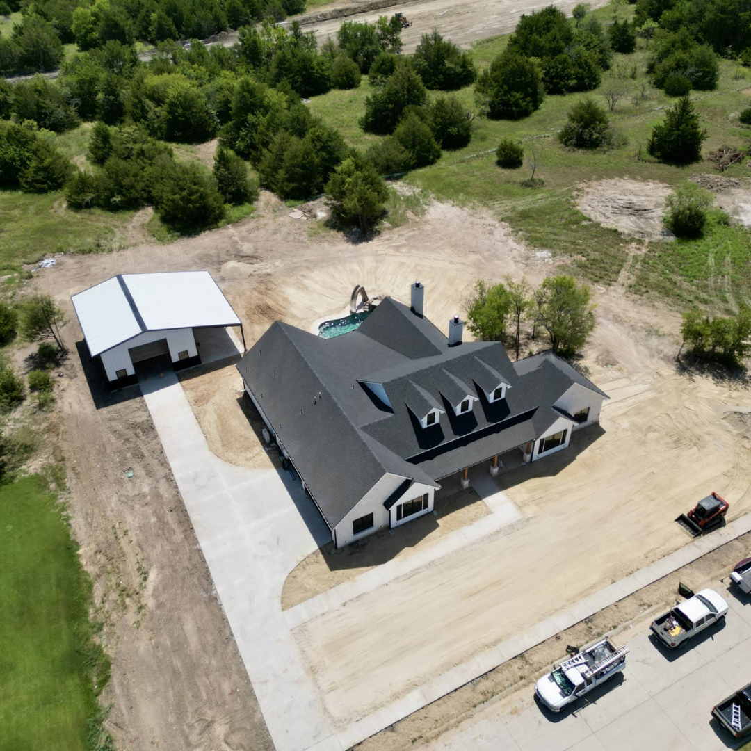 A house under construction with a driveway, a detached garage, and a backyard pool, surrounded by trees and open land.