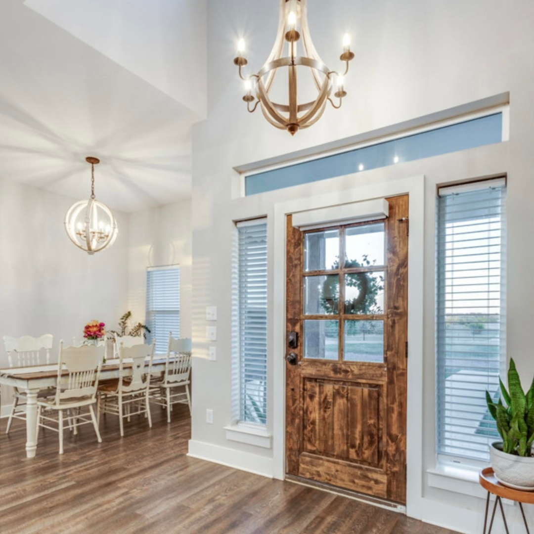 Interior of a house with a wooden front door, two tall windows with blinds, a dining table with chairs, and chandeliers hanging from the ceiling.