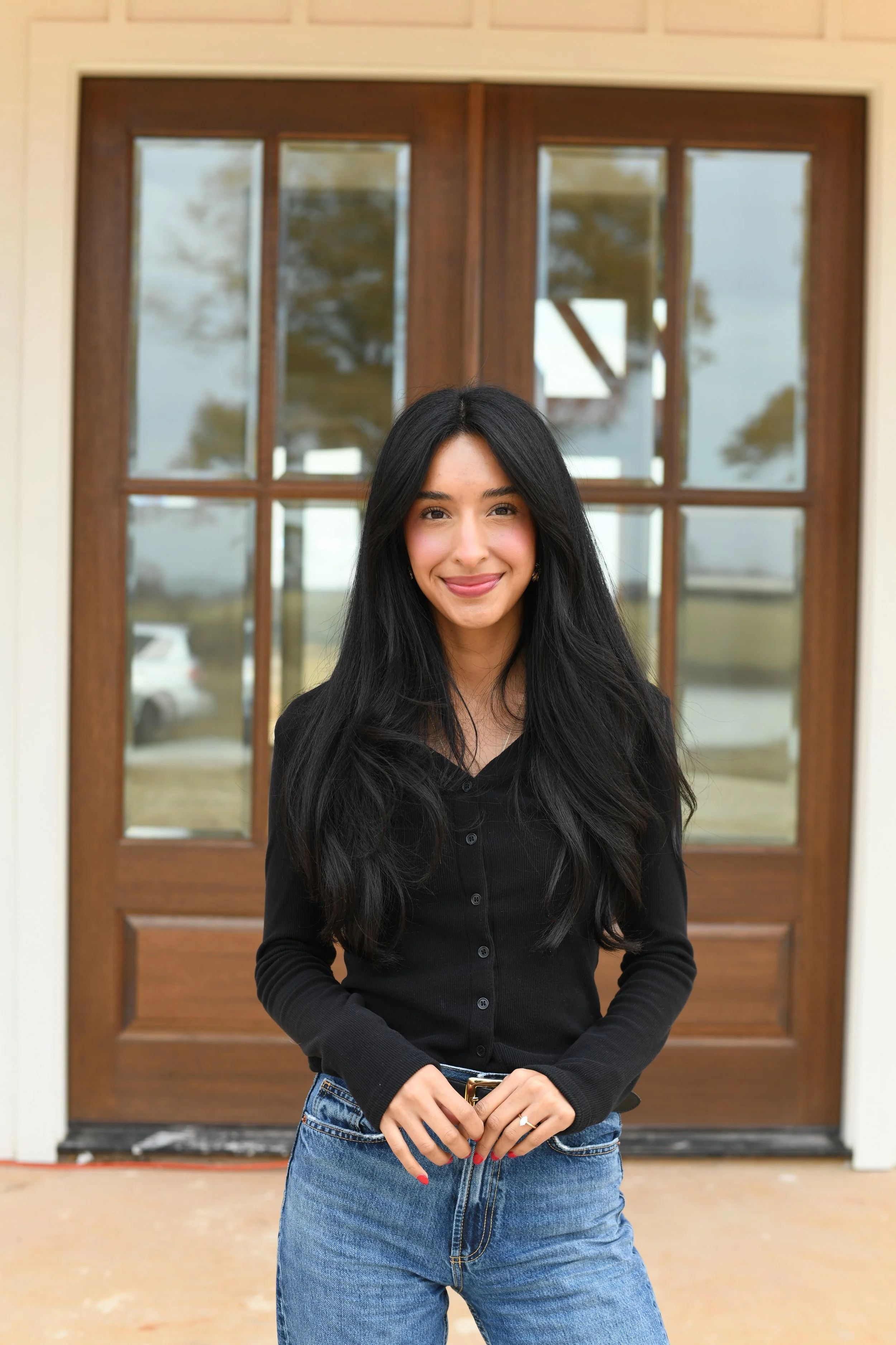 A woman with long black hair, wearing a black button-up shirt and blue jeans, standing in front of a wooden door with glass panels.