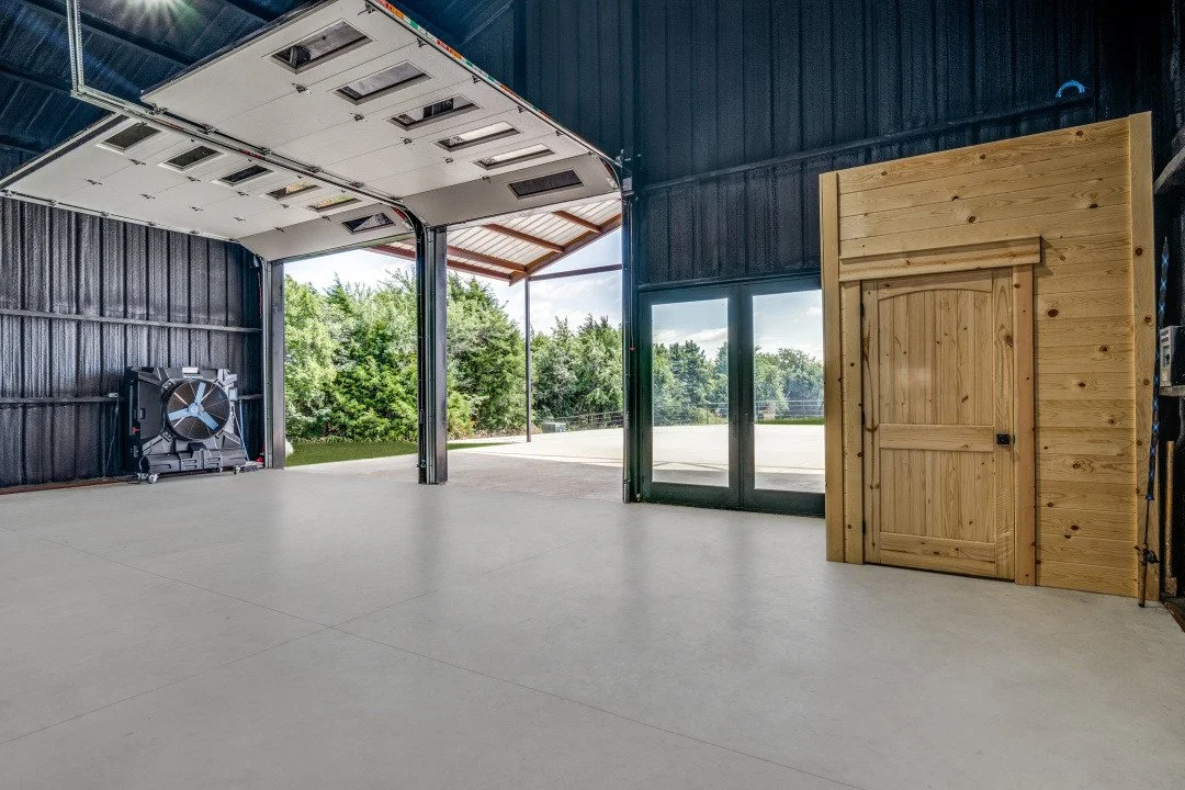 Inside a spacious garage with a large open door, wooden sliding door, and exterior greenery in view.