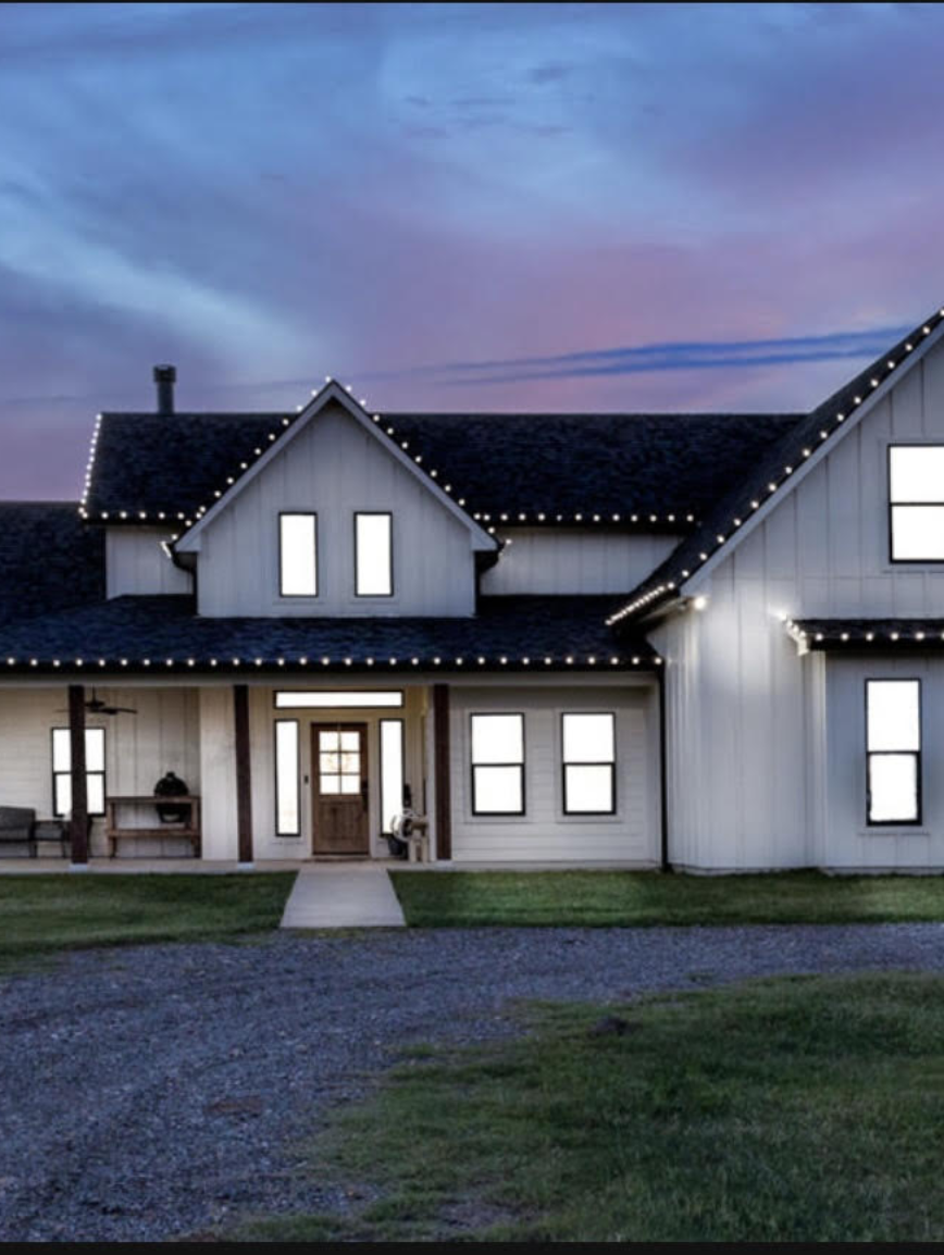 A large white house decorated with Christmas or holiday lights, set against a twilight sky.