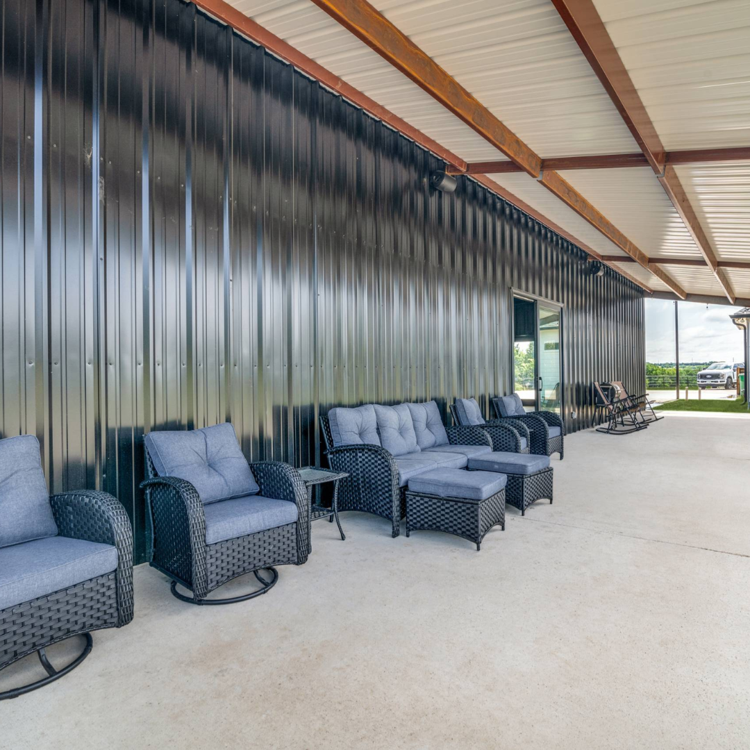 Outdoor patio with wicker armchairs, a wicker loveseat, and a wicker ottoman with light blue cushions, under a metal roof with wooden beams, adjacent to a black metal wall and a door leading inside, with a view of parked vehicles and greenery outside.