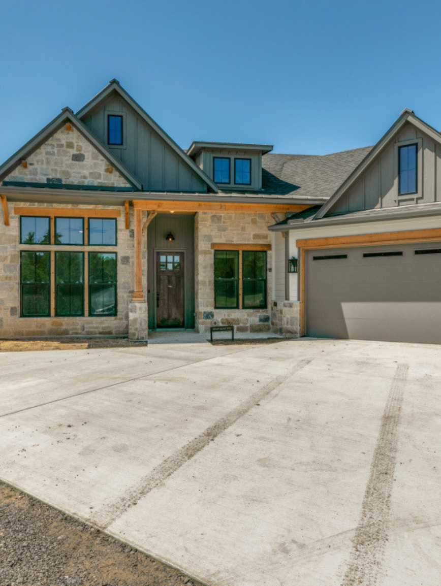 New modern house with stone and gray siding exterior, large windows, and a front driveway with tire marks.