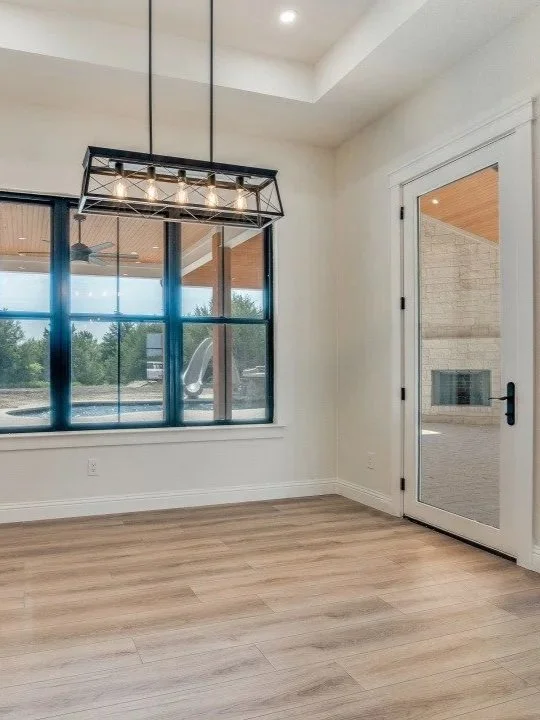 Empty dining or living area with large window, white walls, wood flooring, and a glass door leading outside. Modern rectangular hanging light fixture.