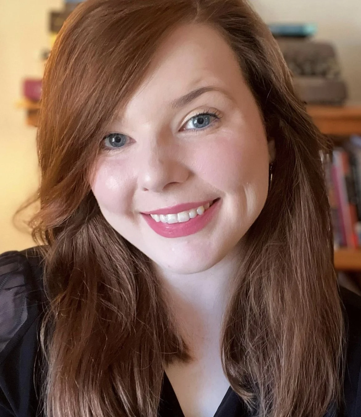 Person with long red hair and blue eyes smiling in a well‑lit professional headshot, with a blurred bookshelf in the background.