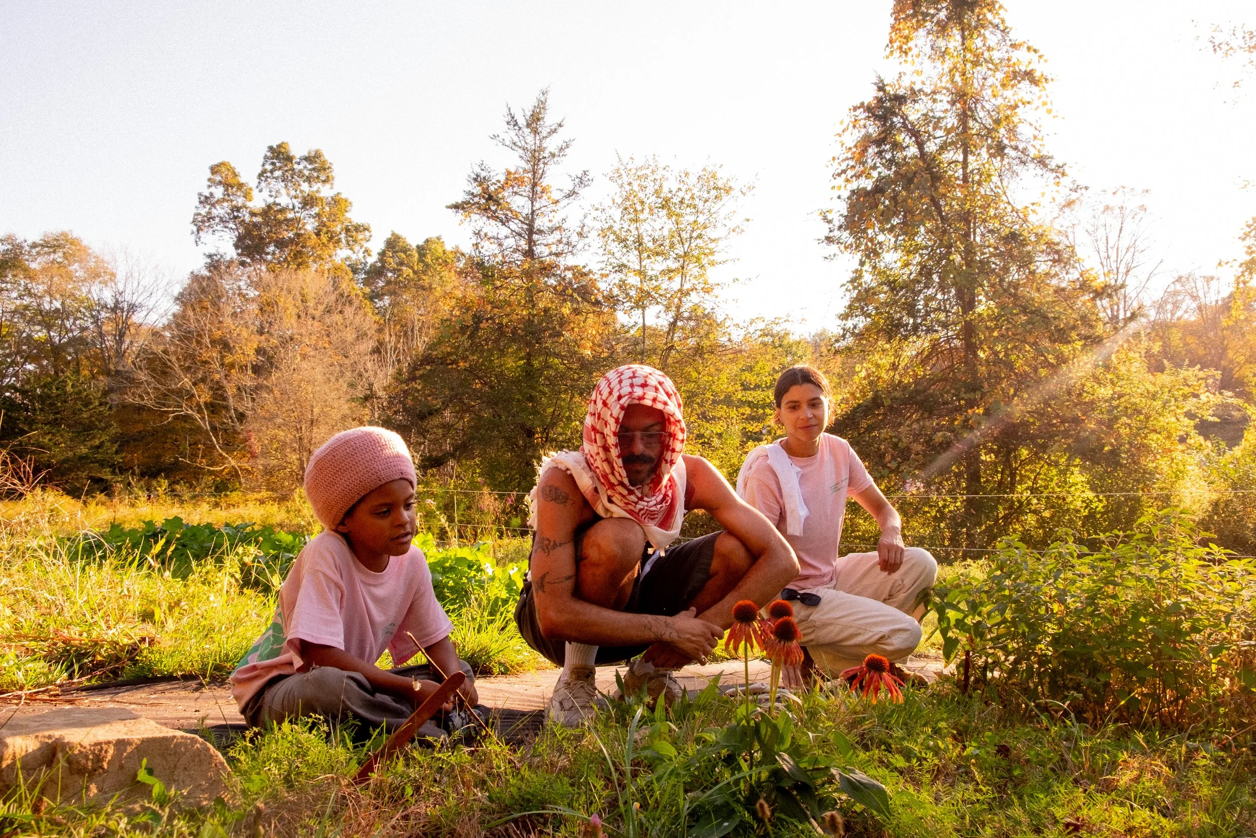 Three people, two children and an adult, are kneeling and sitting on the ground in a garden with flowers and green plants, surrounded by trees in the background during golden hour sunlight.