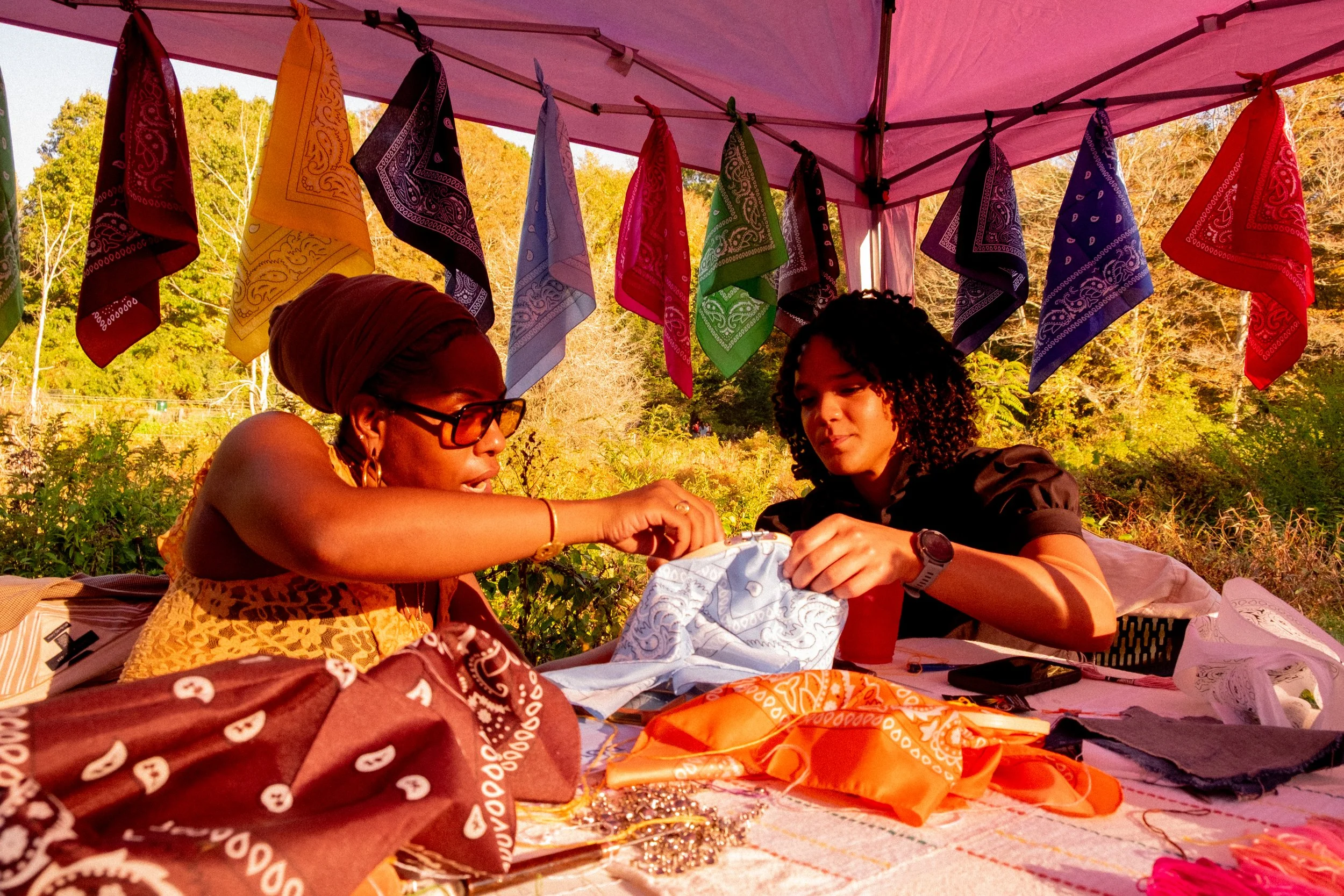 Two women sitting at a table outdoors under a pink canopy with colorful bandanas hanging above, engaged in sewing or crafting, surrounded by greenery and trees in the background.
