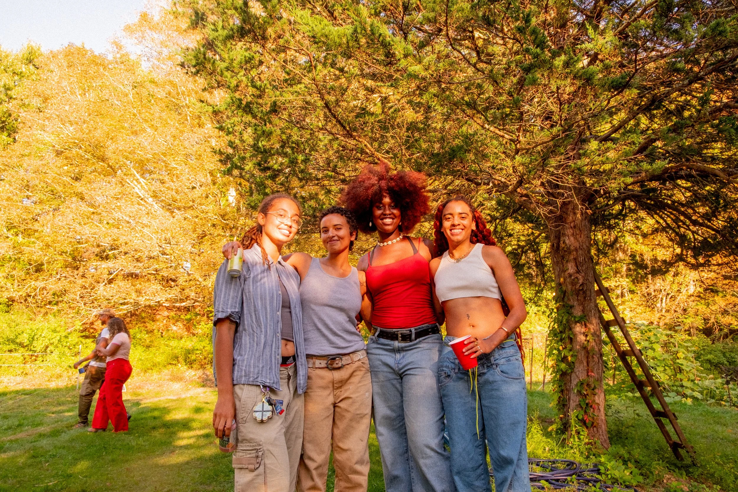 Four women standing together outdoors in a park, smiling and enjoying a sunny day. They are dressed casually, with two holding drinks, and are positioned in front of a large tree with green foliage, with couples in the background.