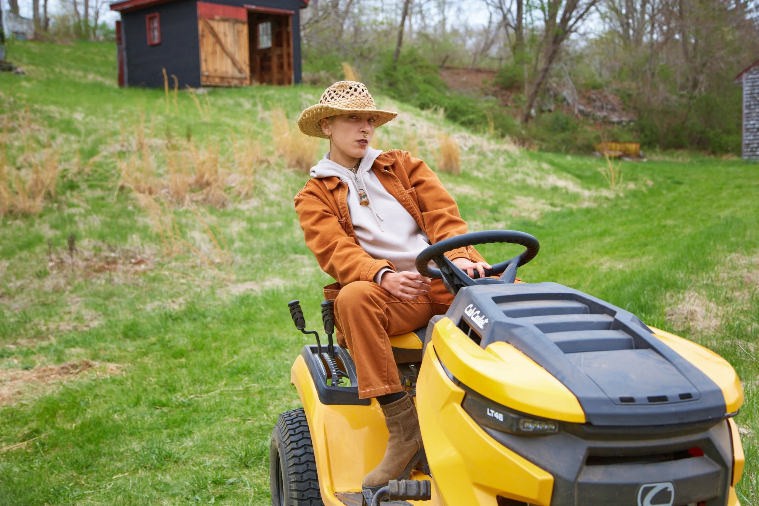 A woman wearing a wide-brimmed straw hat, a brown jacket, a white hoodie, and brown pants sitting on a yellow ride-on lawn mower in a grassy backyard.