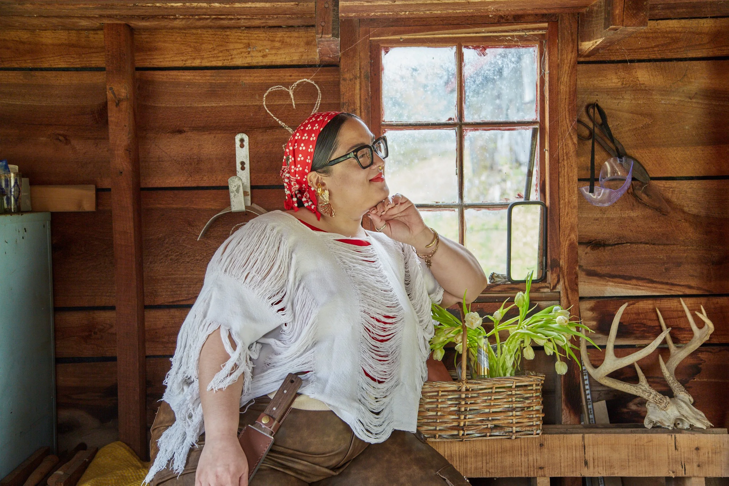 Woman with glasses and a red bandana in a wooden room, looking thoughtfully by a window with a heart drawn above her head.