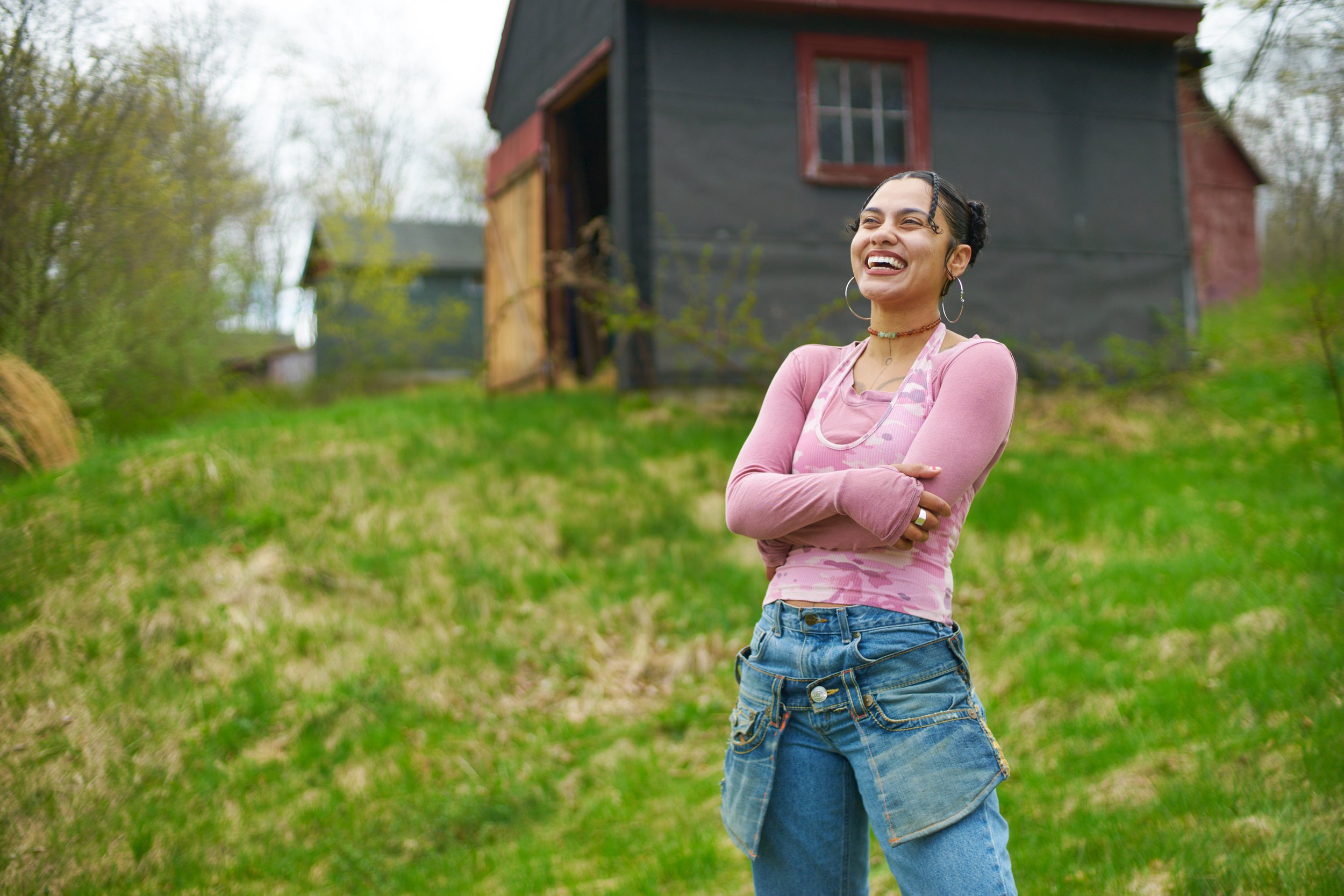 A woman standing outdoors on a grassy hill, smiling and laughing with arms crossed, in front of a black and red barn-like structure.