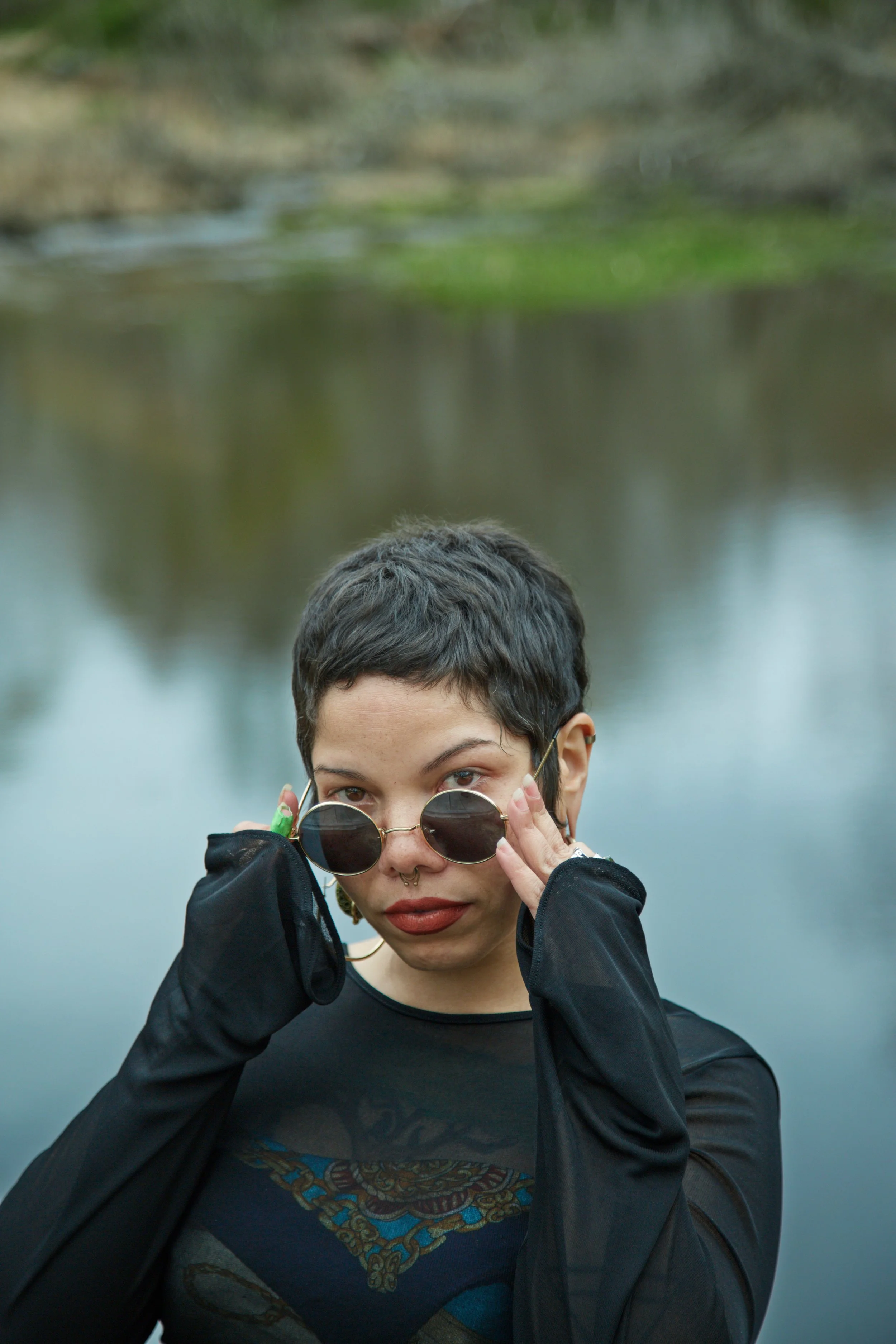 A person with short black hair and sunglasses standing outdoors near a body of water, adjusting their sunglasses with both hands.