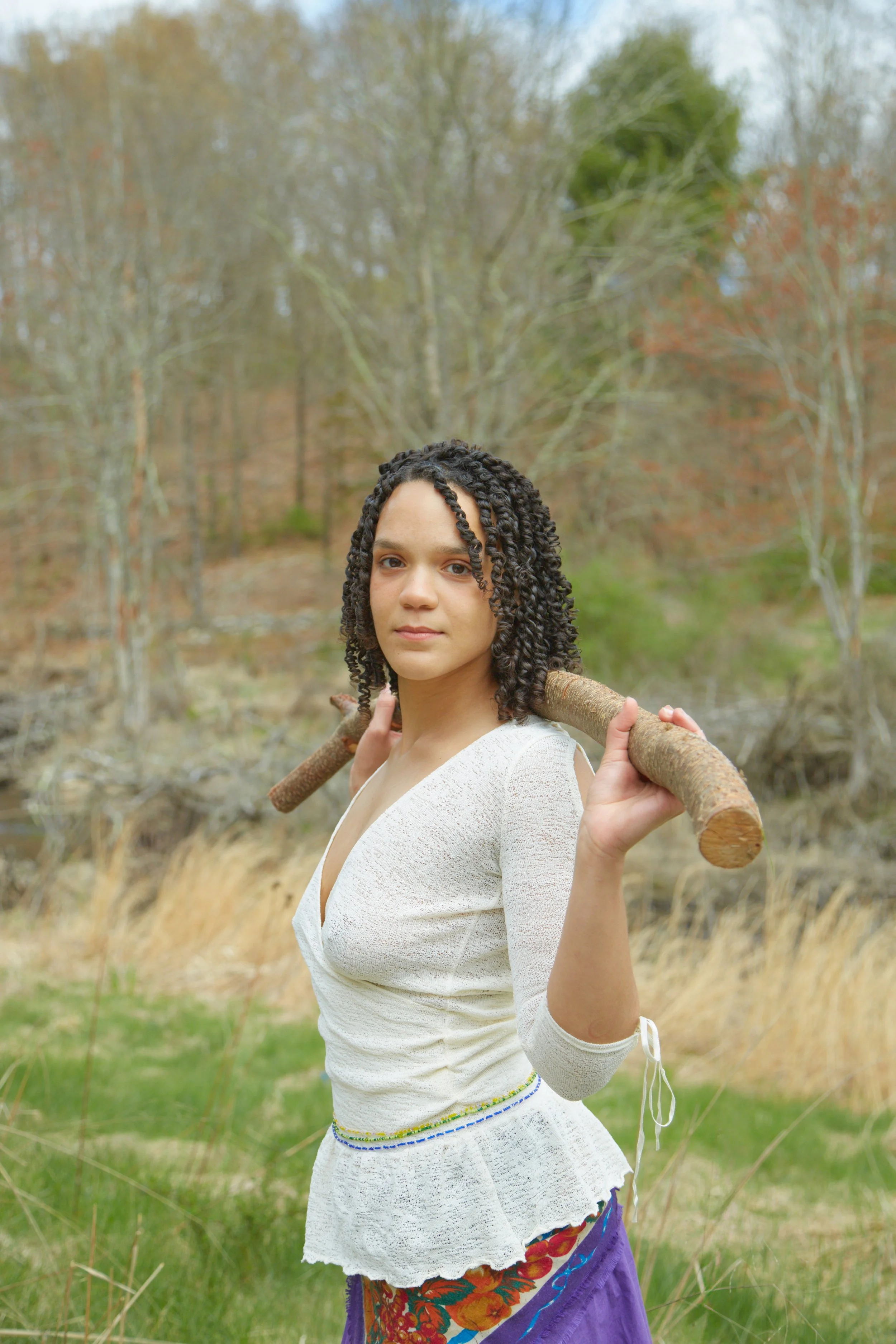 Young woman with curly hair holding a stick across her shoulders outdoors in a natural setting with trees and grass.