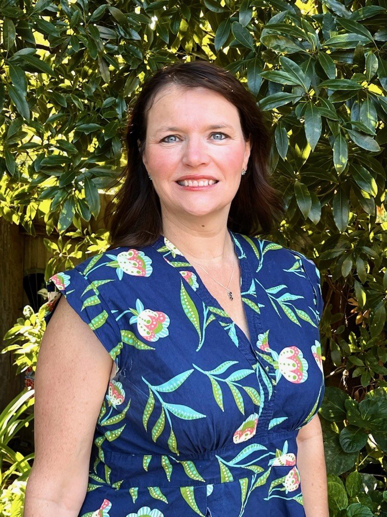 Portrait of a woman with shoulder-length brown hair and blue eyes, wearing a blue floral dress, standing outdoors in front of green foliage.