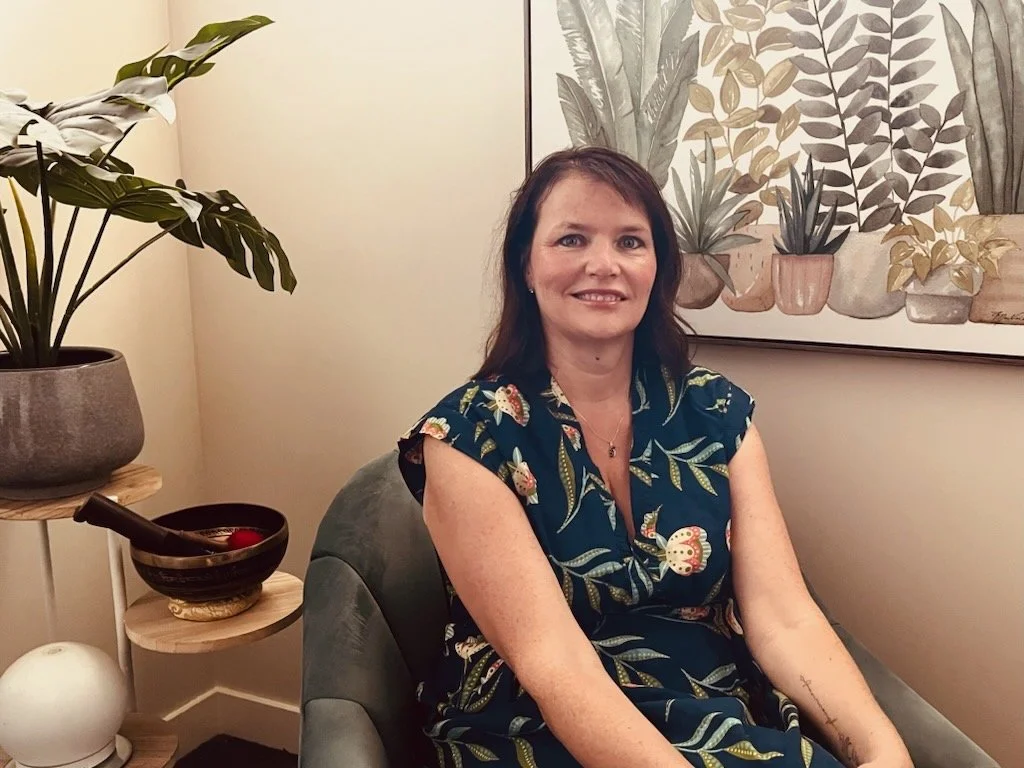 A smiling woman with shoulder-length brown hair, wearing a dark floral dress, sitting in a living room next to a framed botanical print on the wall, with a large potted plant and decorative bowls on a side table.