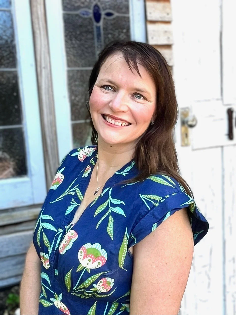 A woman with shoulder-length dark brown hair and blue eyes, smiling, wearing a blue floral dress with pink and green flowers, standing outside in front of a white wooden door with a decorative window.