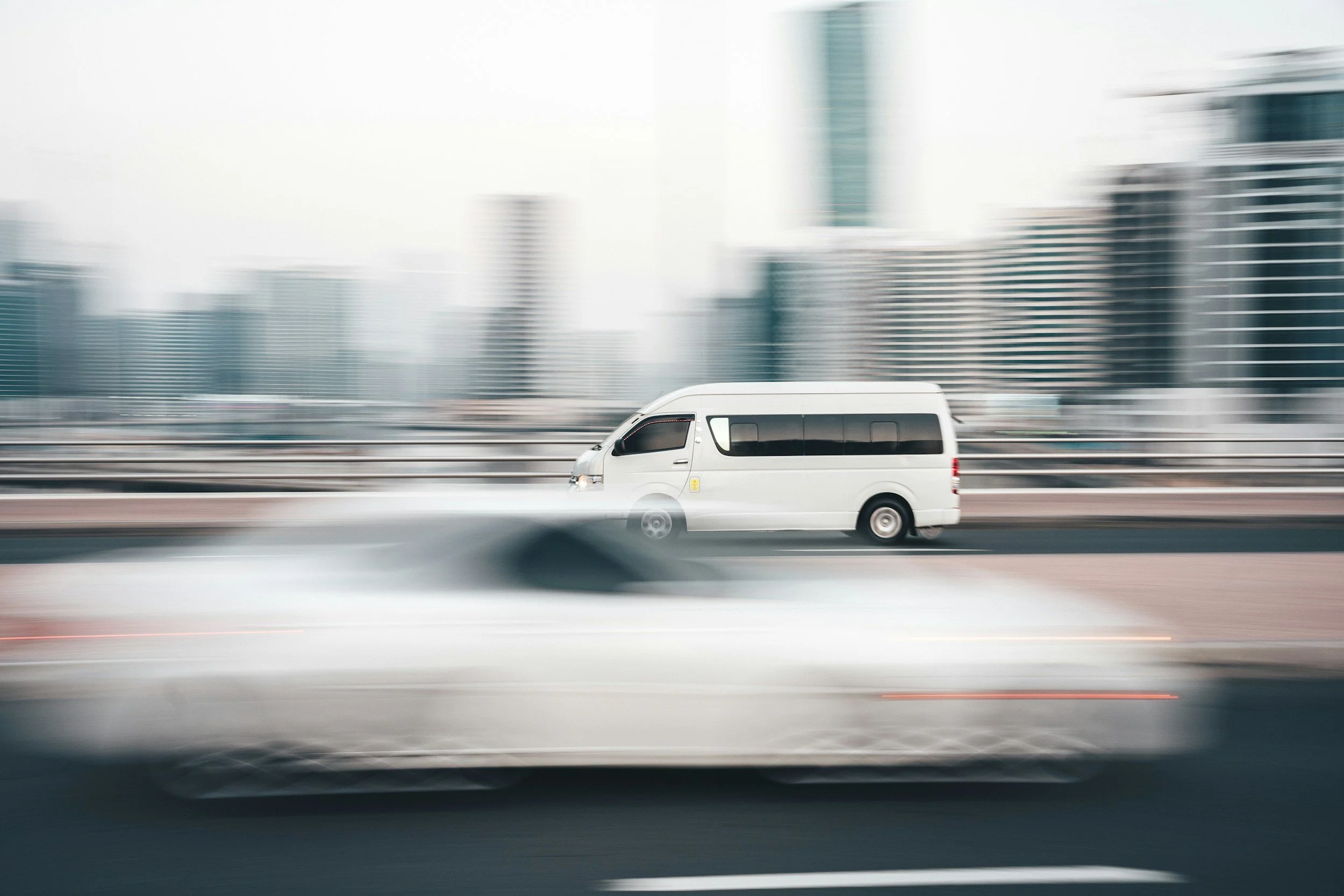 A white passenger van driving on a highway in an urban area with high-rise buildings in the background.