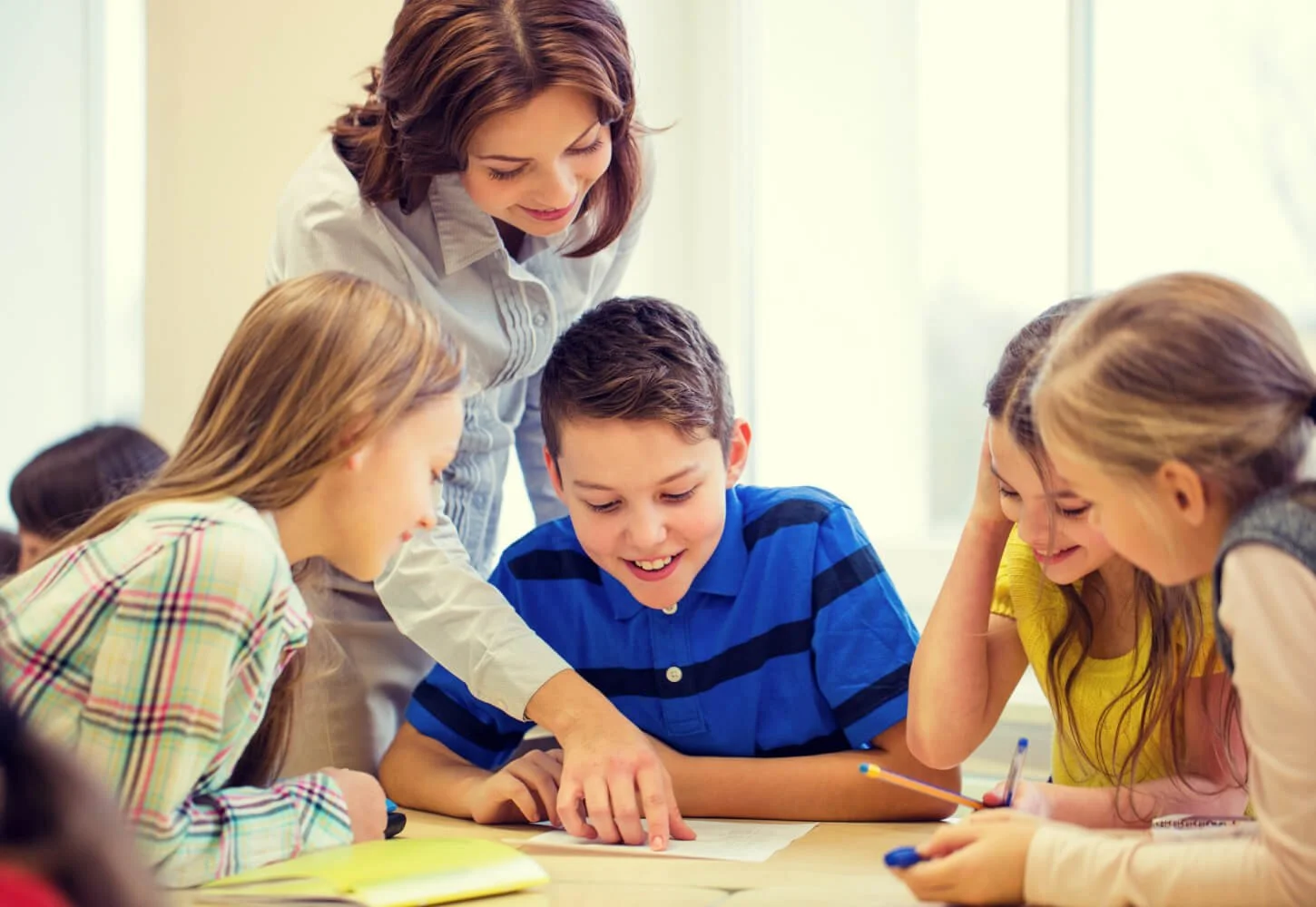 A teacher assisting students during a classroom activity or lesson, with students smiling and engaging with papers and pencils.