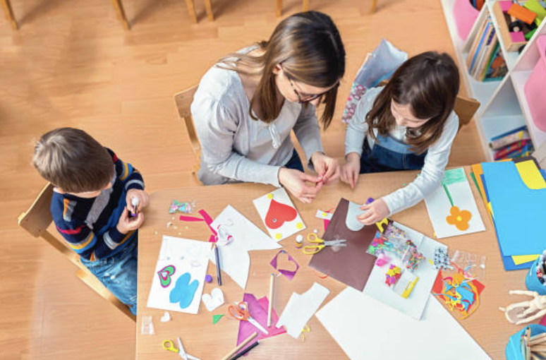 A woman and two children engaging in arts and crafts at a wooden table, with paper, scissors, and colorful cut-out shapes.