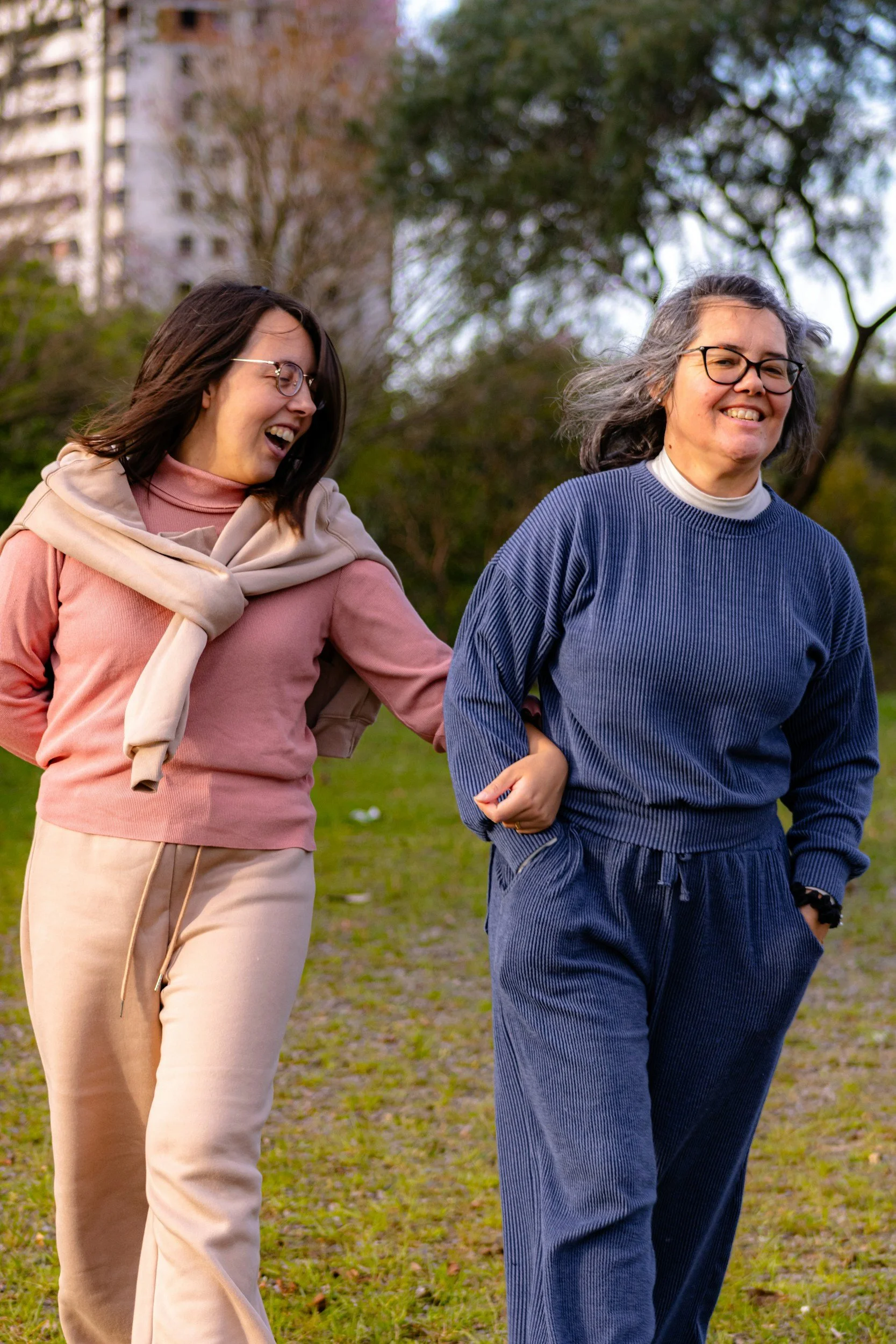 Two people walking through a grassy park with their arms linked.