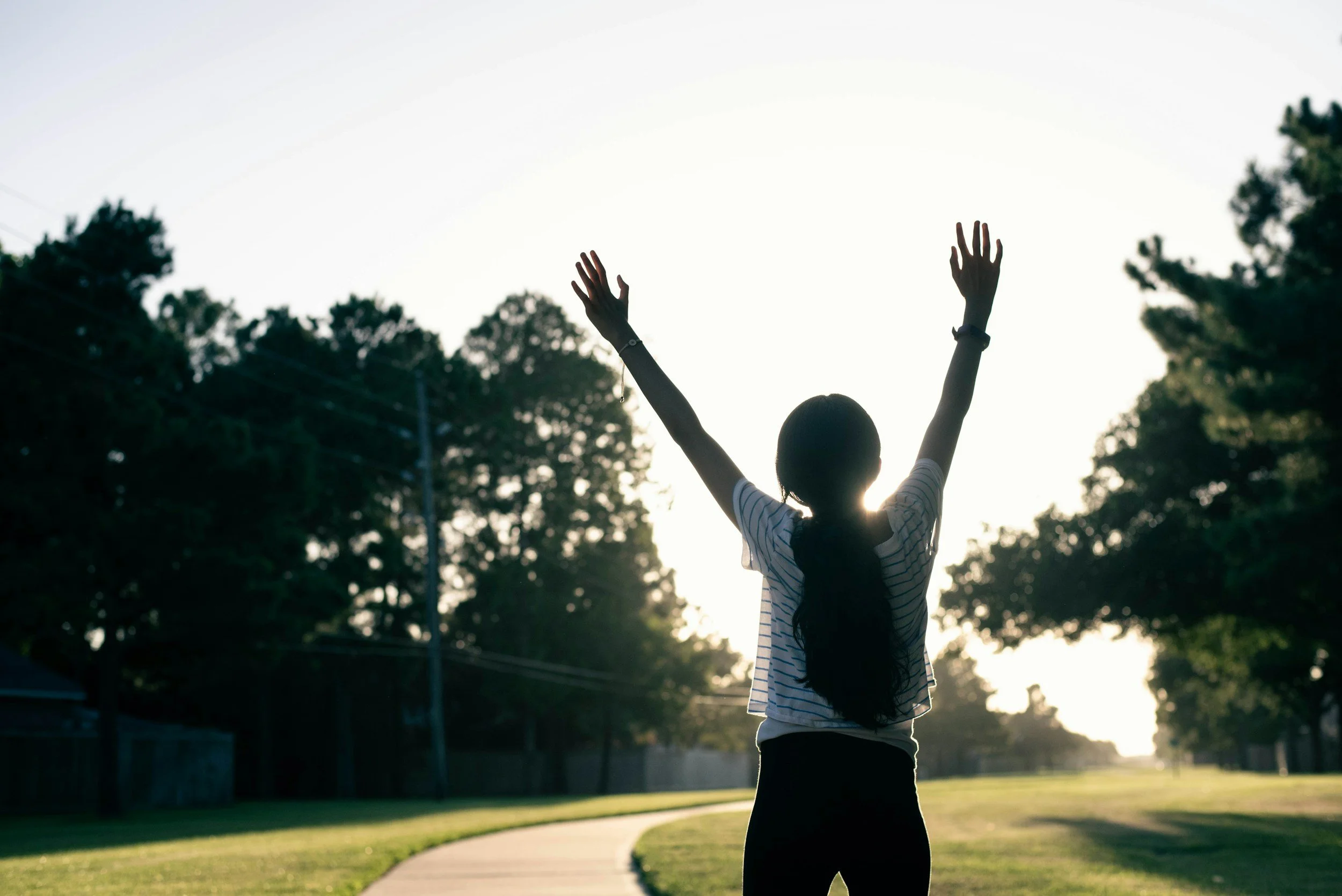 Person standing outside with their hands in the air in celebration, their back to the camera. Wellness, health, mental health, self-care, connection, therapy, holistic