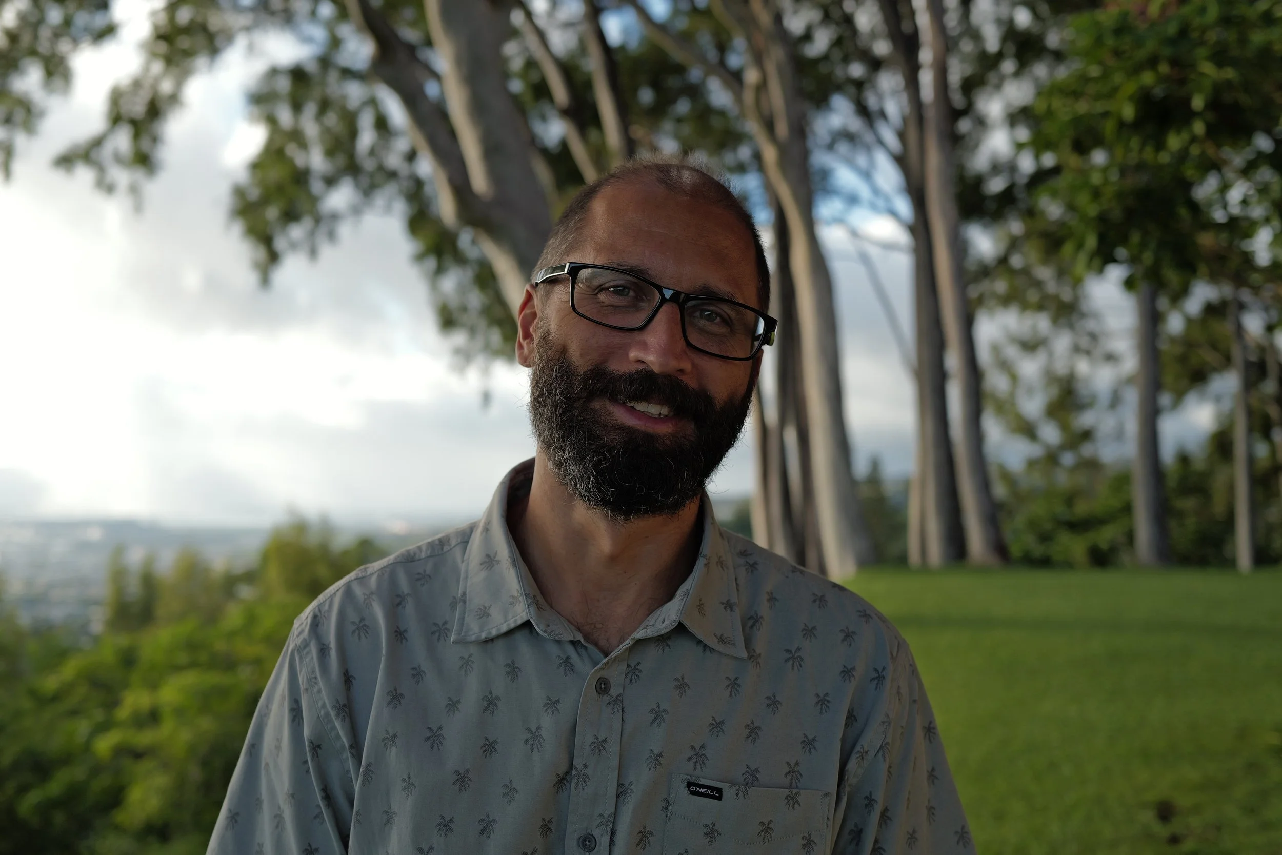 Fabio Cardoso smiling outdoors in front of trees and a cloudy sky. Oahu Family Therapy, Honolulu, HI. Providing Individual, Couples, Pre-marital, and Family therapy.