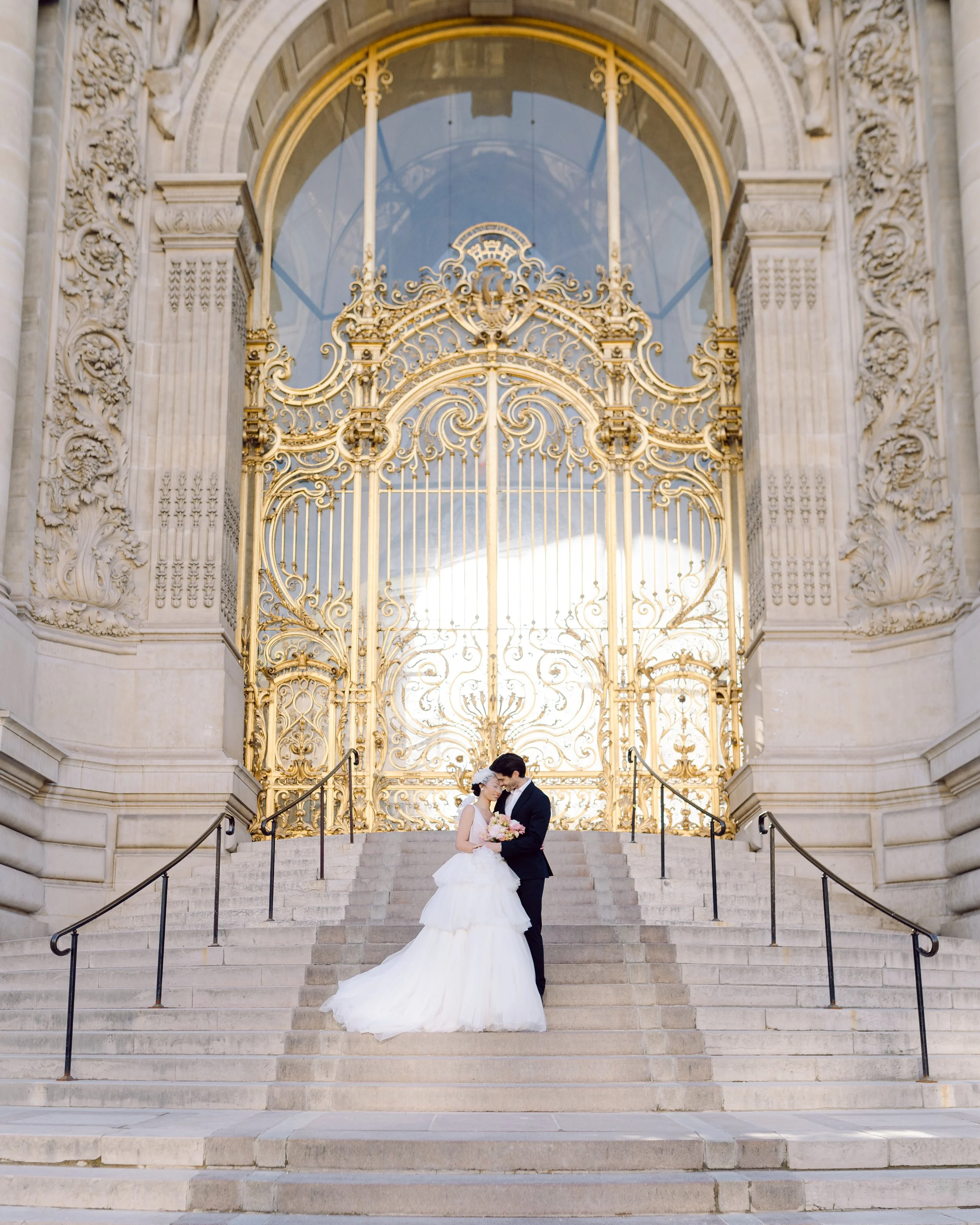 Un couple de mariés debout sur des escaliers en pierre devant une grille dorée ornée, à l'extérieur d'un bâtiment historique en pierre.