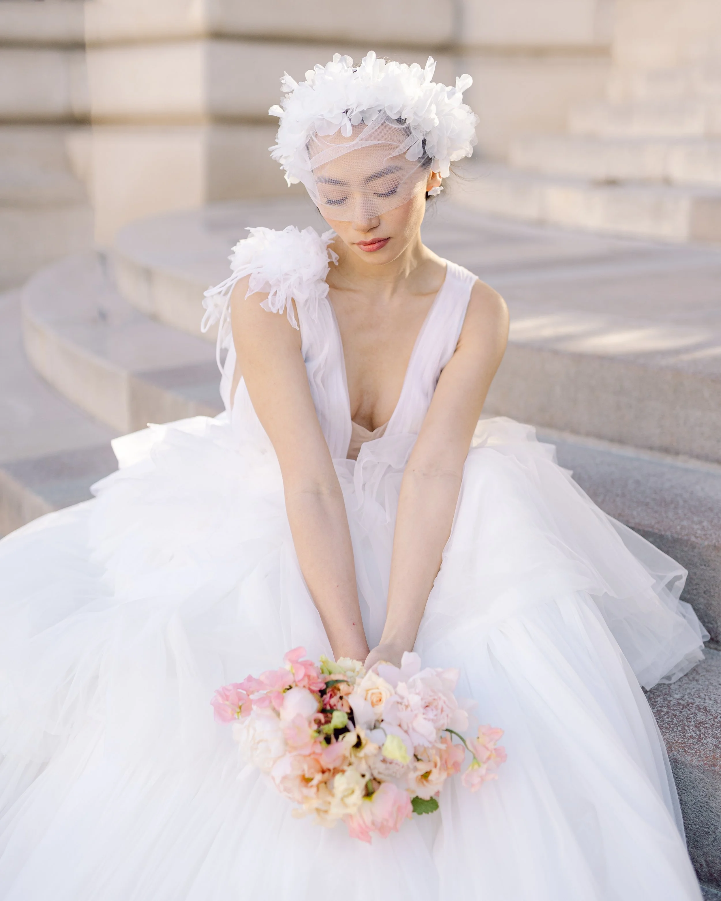 Une femme habillée en robe de mariée blanche, portant un voile et un chapeau de fleurs, assise sur des marches en pierre, tenant un bouquet de fleurs.