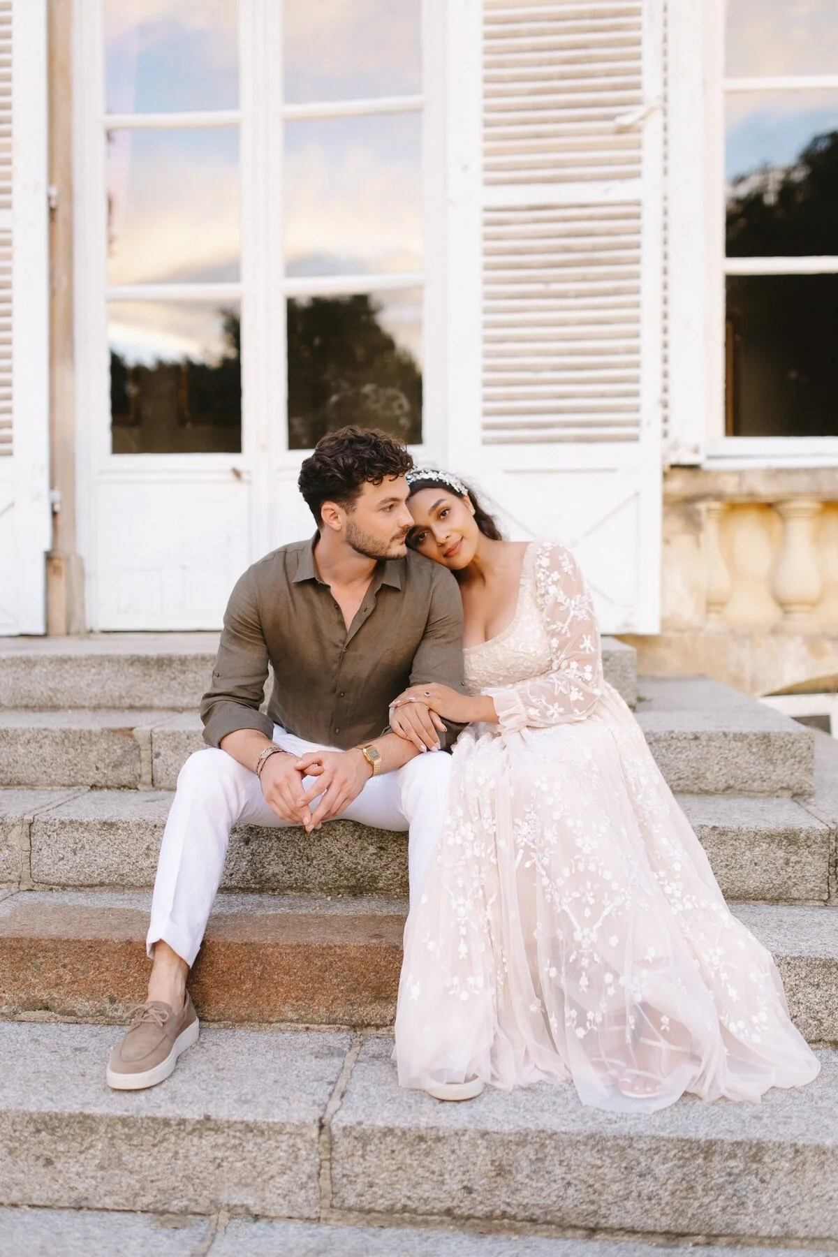 Un couple assis sur des escaliers en pierre devant une grande fenêtre avec volets blancs, lors du coucher de soleil.