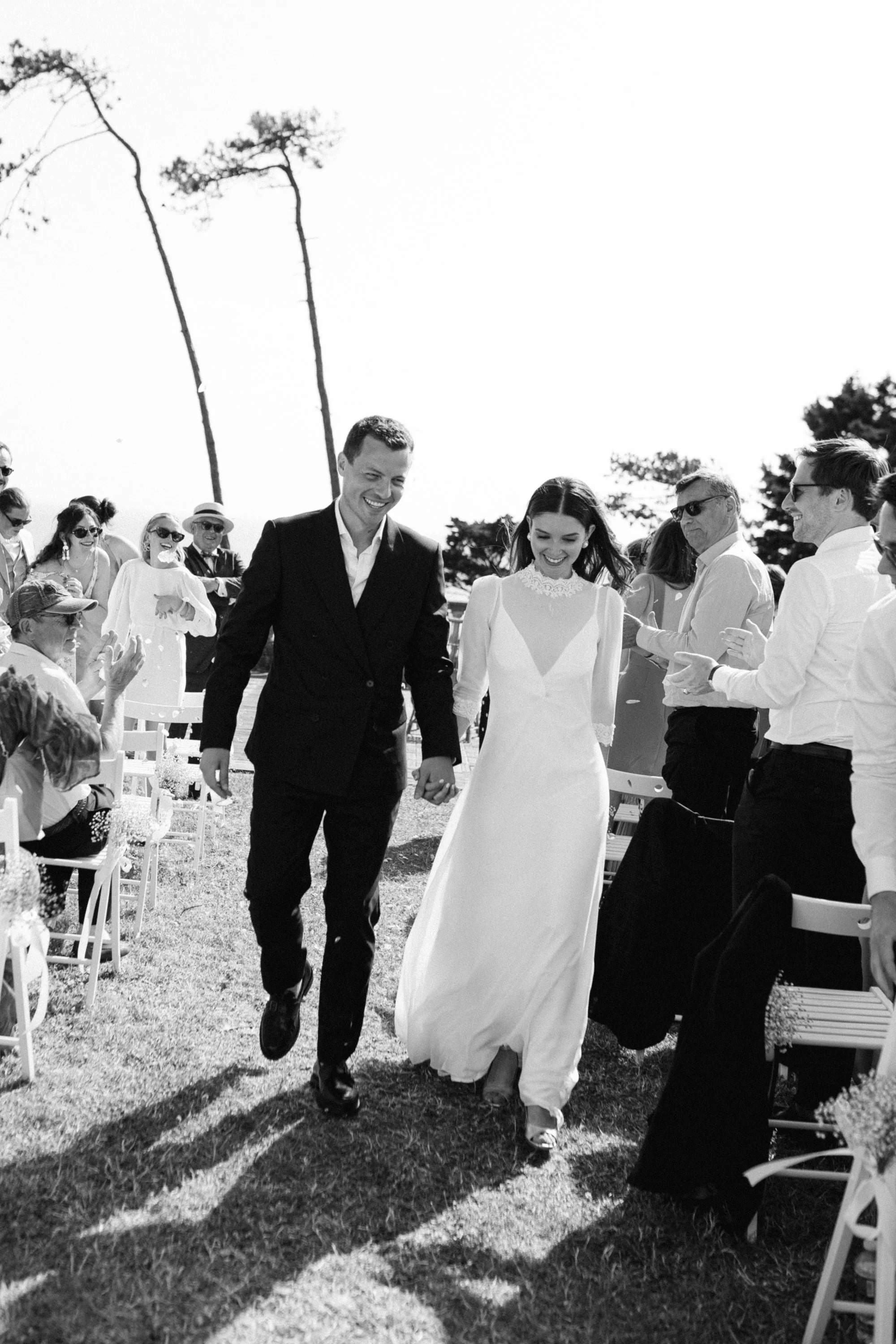 Un couple en costume de mariage marche main dans la main lors de leur cérémonie, entouré de invités assis ou debout en plein air, avec des arbres en arrière-plan.