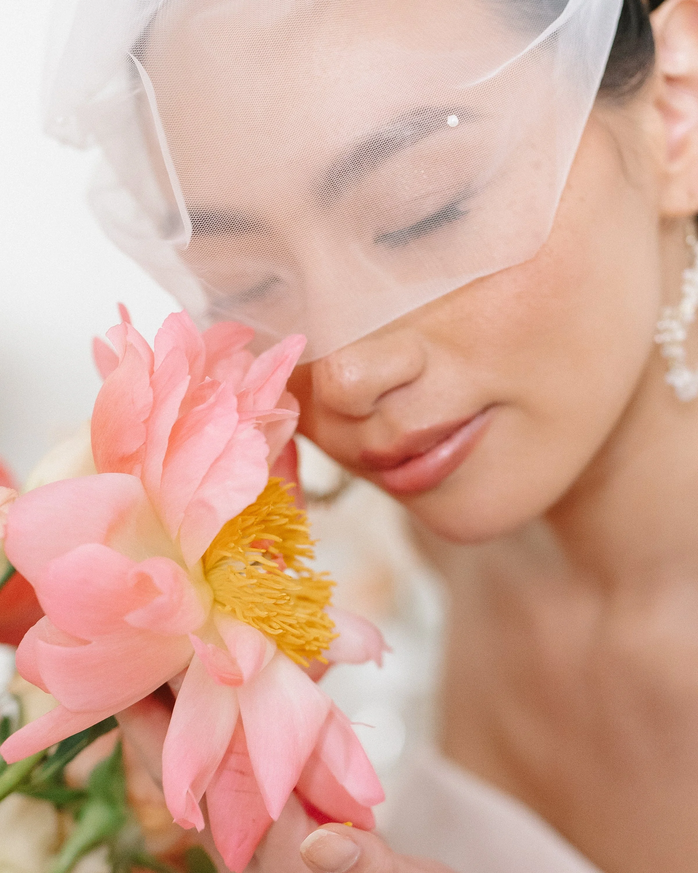 Une femme portant un voile de mariée blanc tient un grand bouquet de fleurs roses, son visage est en gros plan, elle a les yeux fermés et un sourire doux.